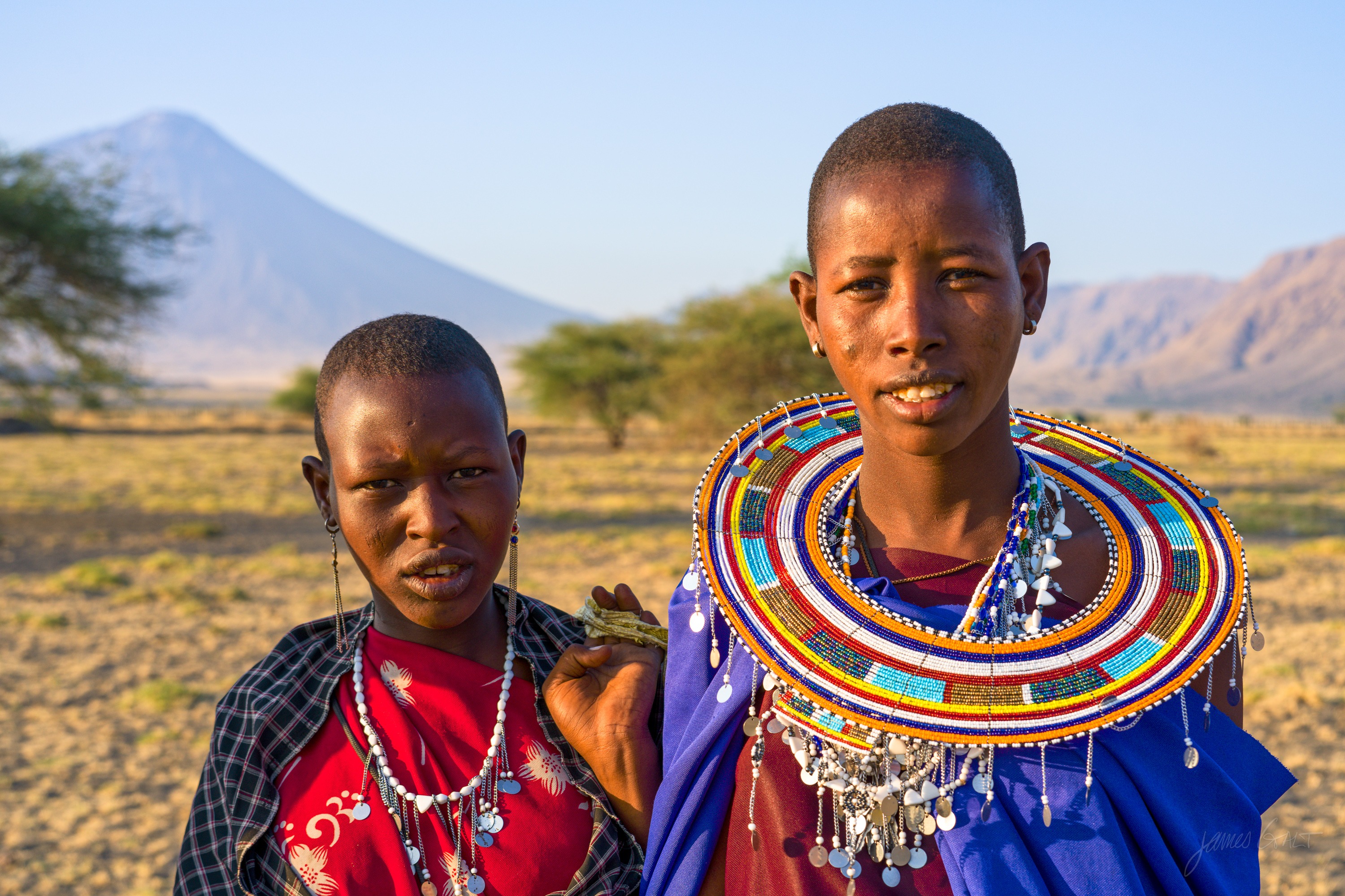 The same two women in colour, in red robes and layered blue, red and yellow bead collars, the grassland and a distant volcano warm in the morning light.