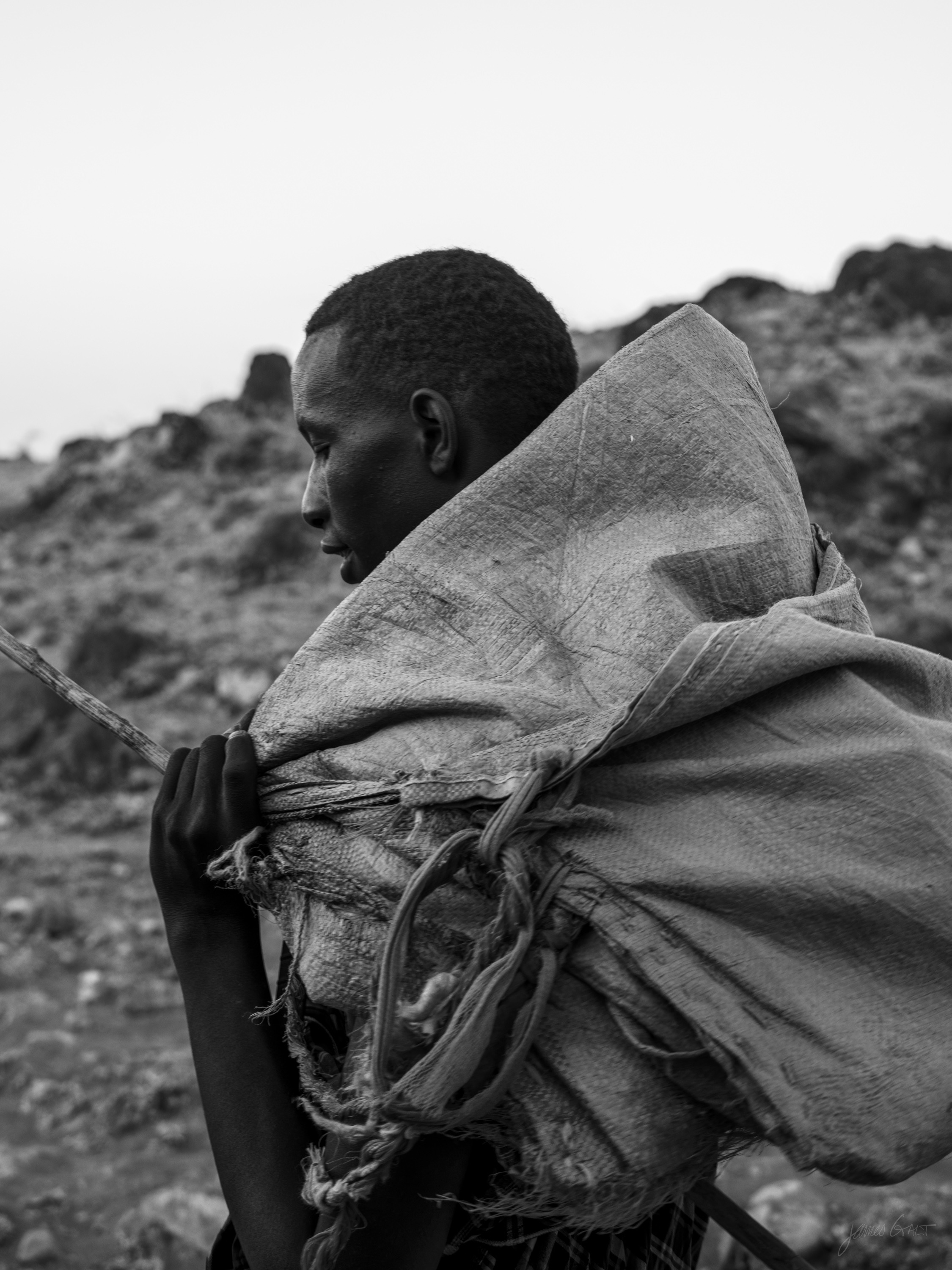 Black and white photograph of a person seen in profile, wrapped tightly in a blanket over the head and shoulders, standing against a bare rocky slope at dusk.