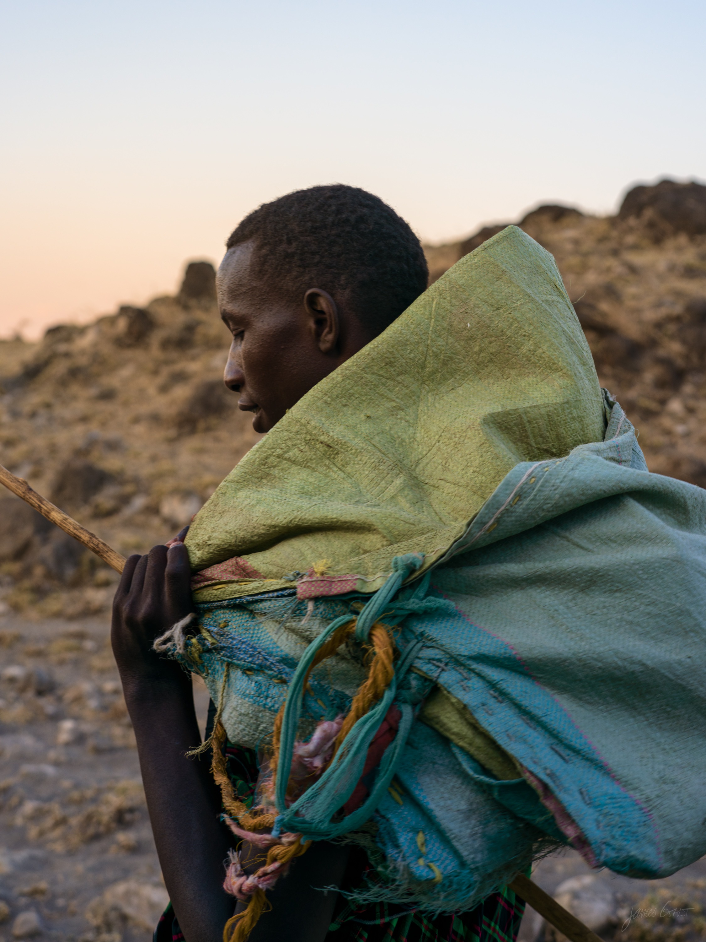 The same figure in colour, wrapped in a green and blue shuka pulled up over the head, standing in profile against a hillside with warm dusk light along the horizon.