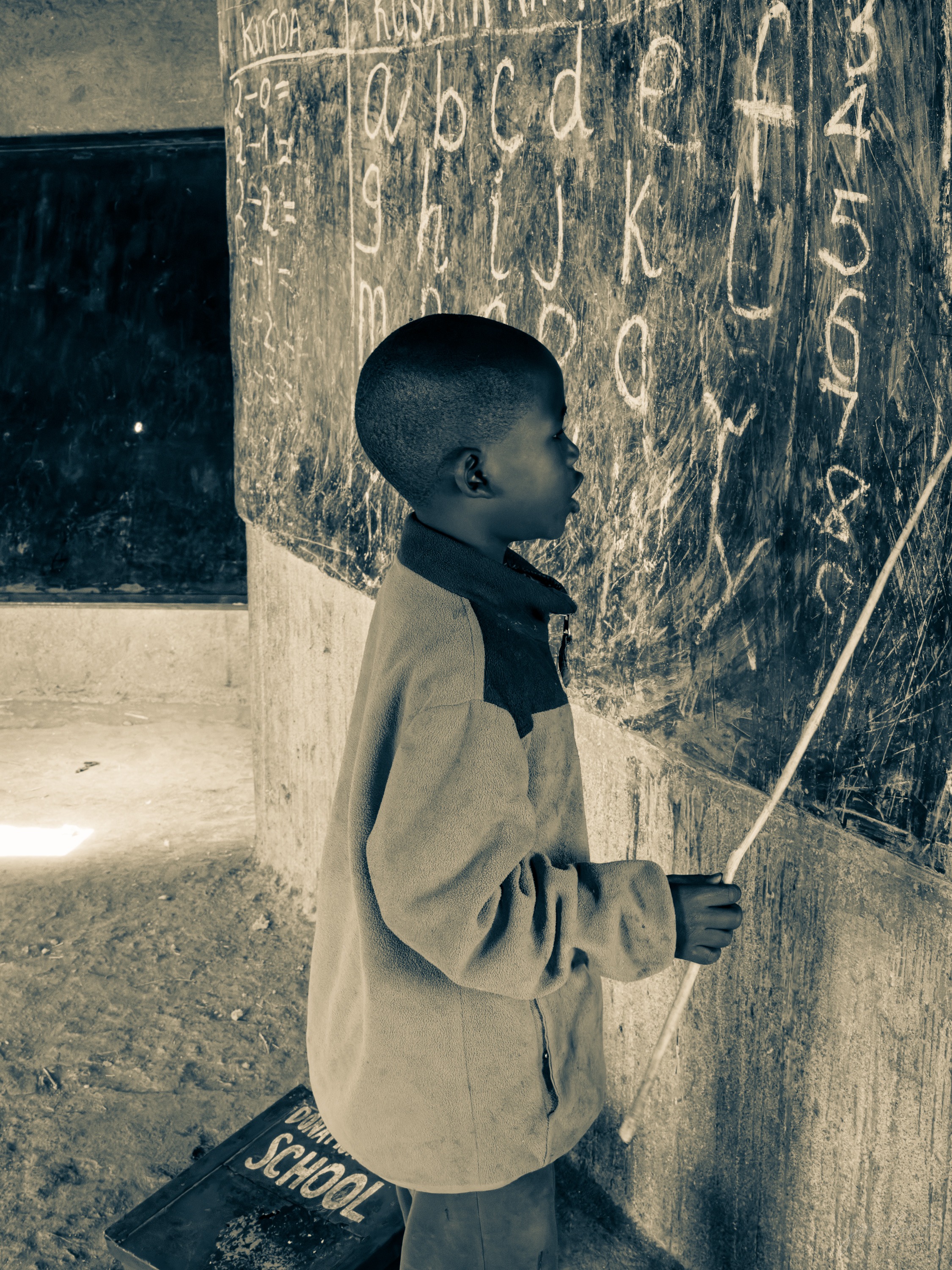 Black and white photograph of a young boy standing at a chalkboard covered in handwriting, holding a stick pointer, in a dim classroom.