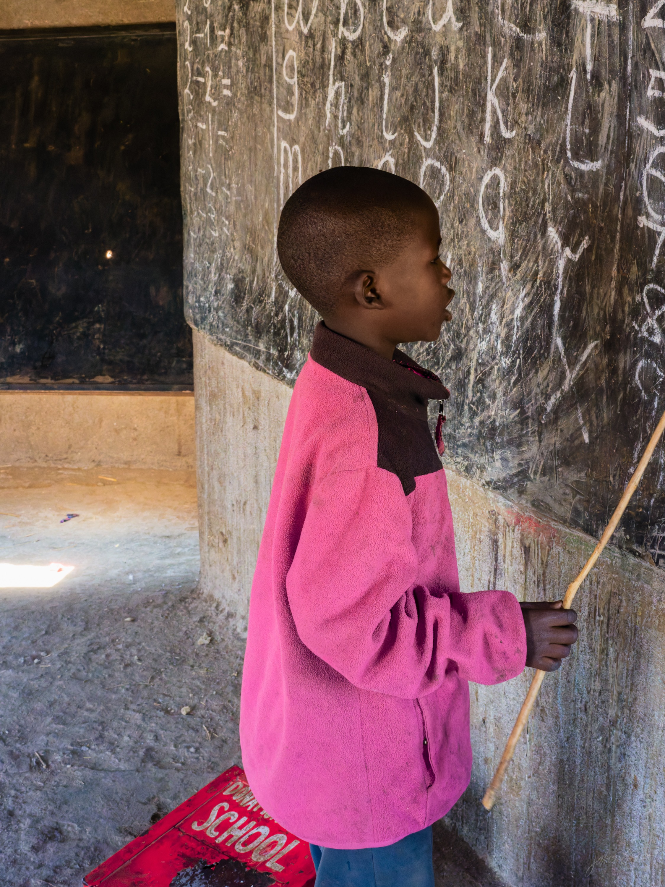 The same boy in colour, in a bright pink jacket, at a chalkboard full of white writing, warm light from a doorway falling on him.