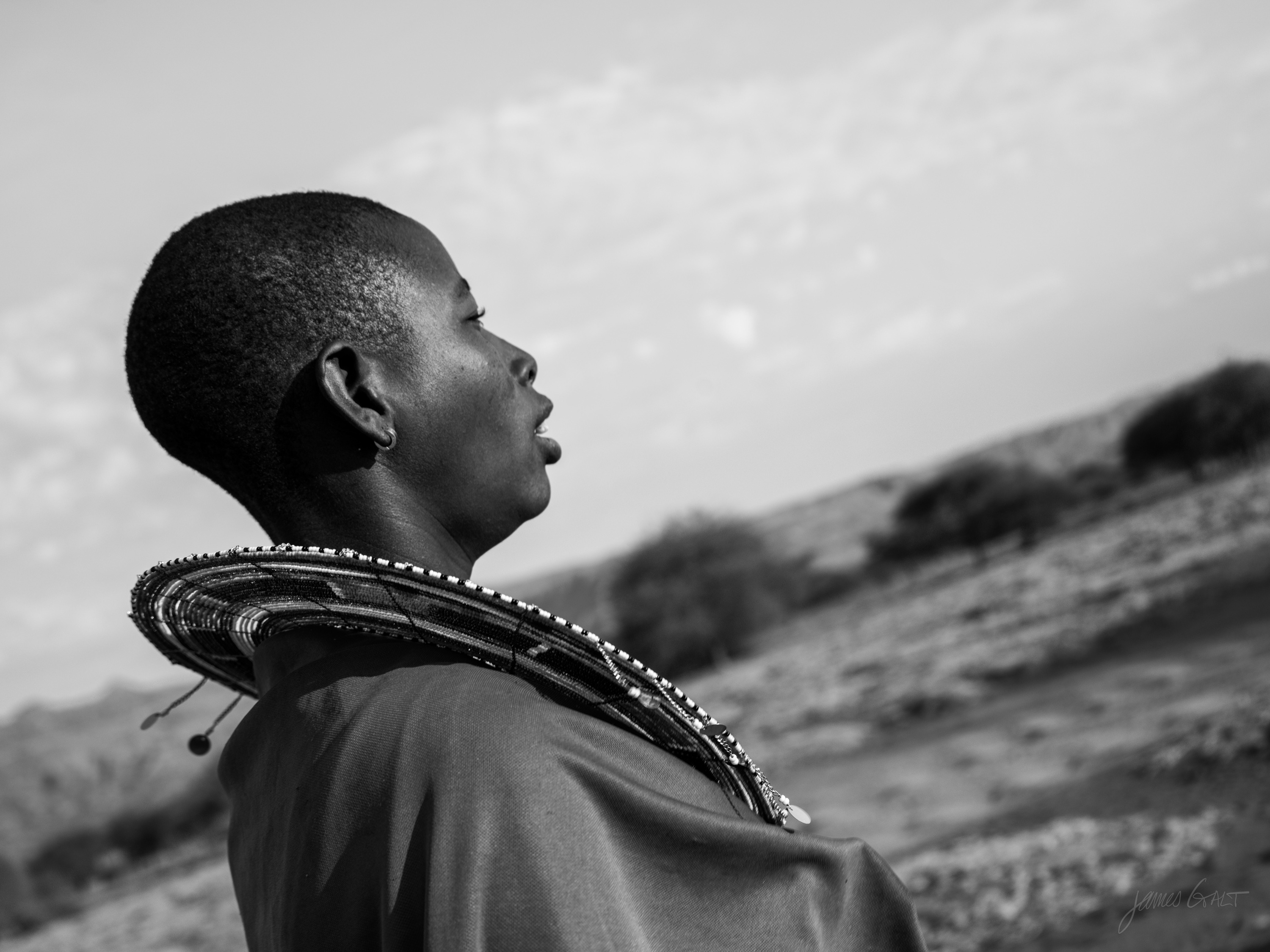 Black and white profile of a Maasai woman with a shaved head and large beaded earrings, looking upward, a beaded collar at her neck, open sky behind.
