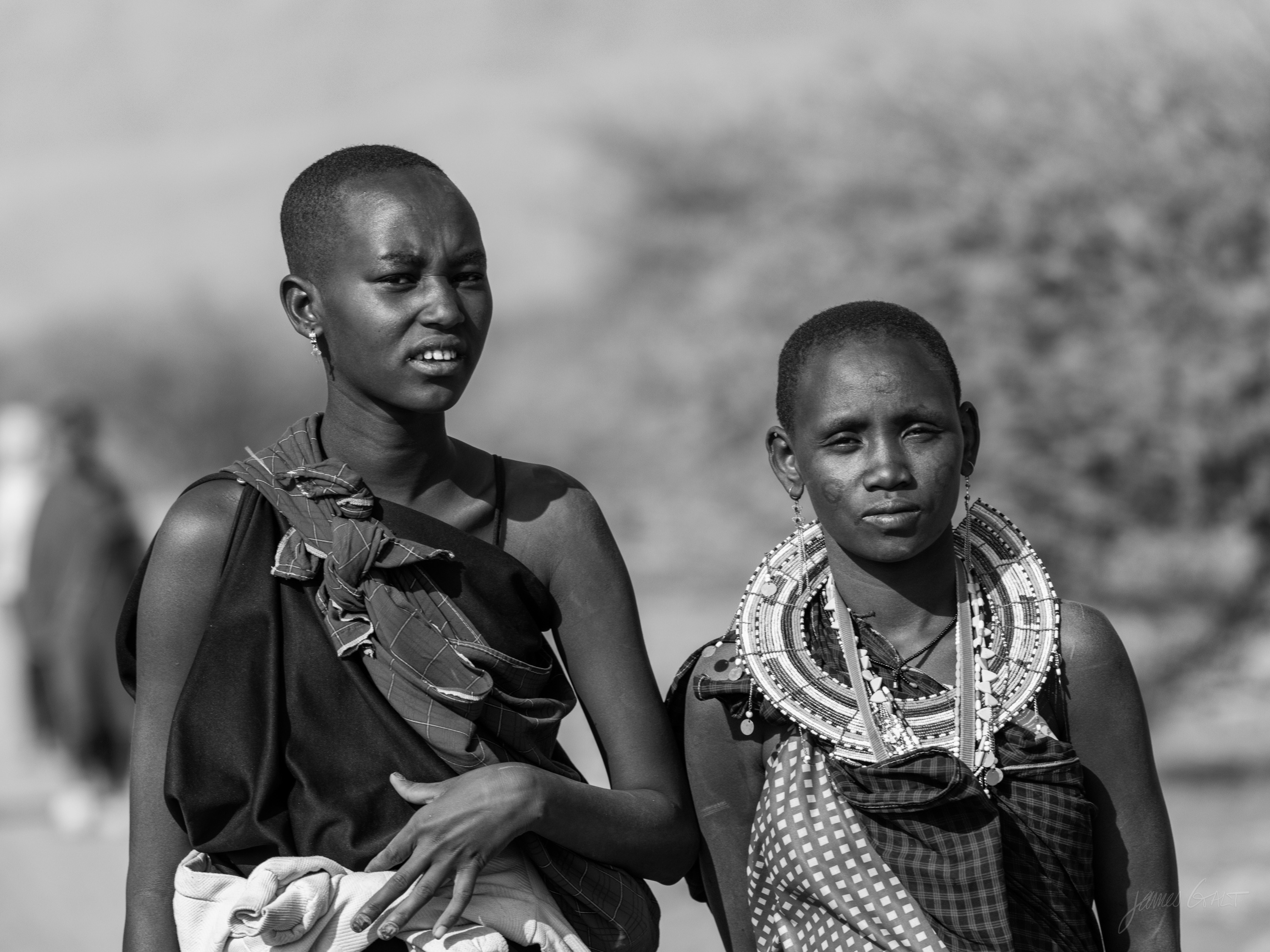Black and white photograph of two Maasai women outdoors, one turned toward the other, both in beaded collars, soft savannah behind them.