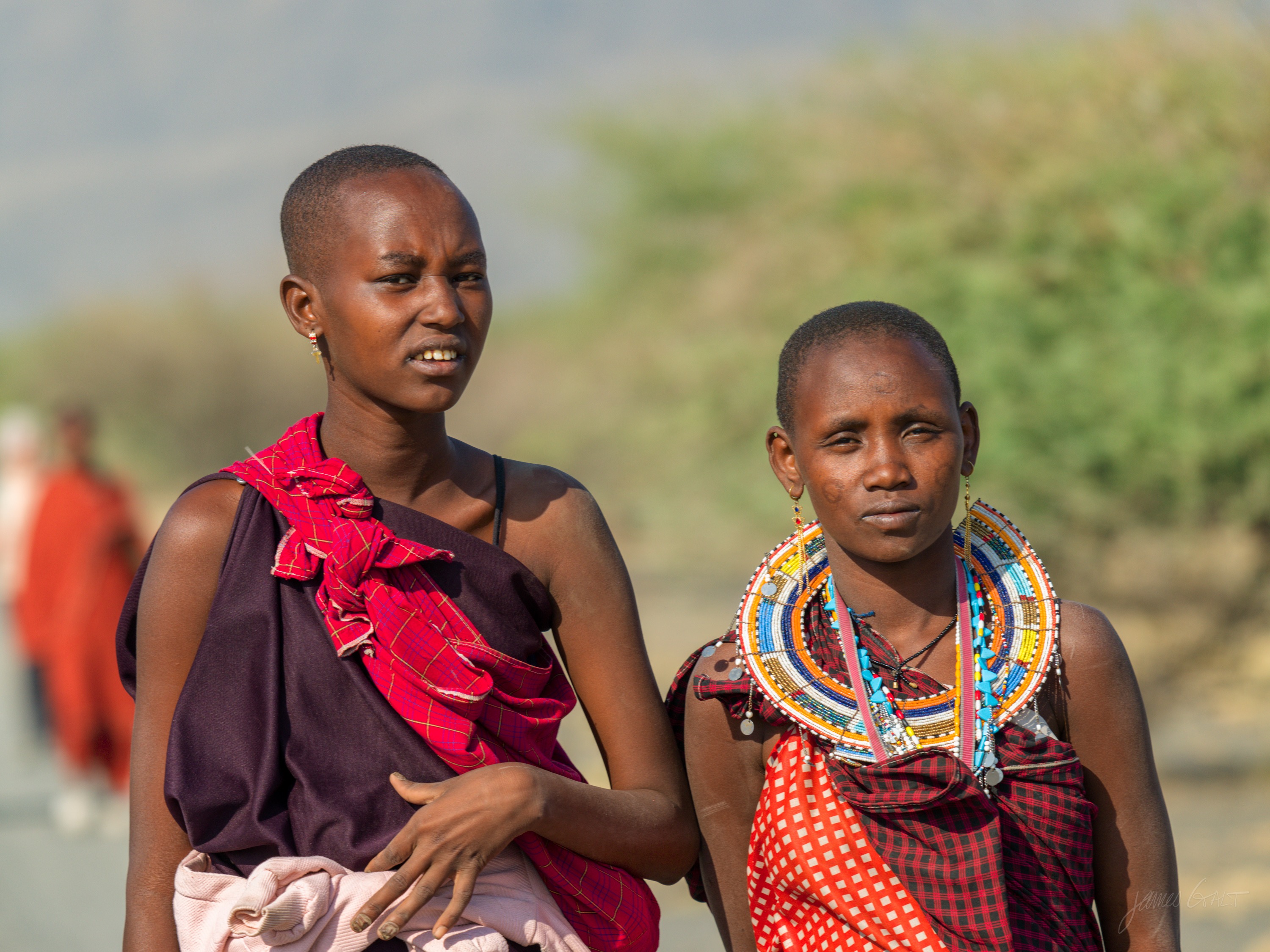 The same two women in colour, one in maroon and one in red with a multicoloured bead collar, green and gold bush behind them in morning light.