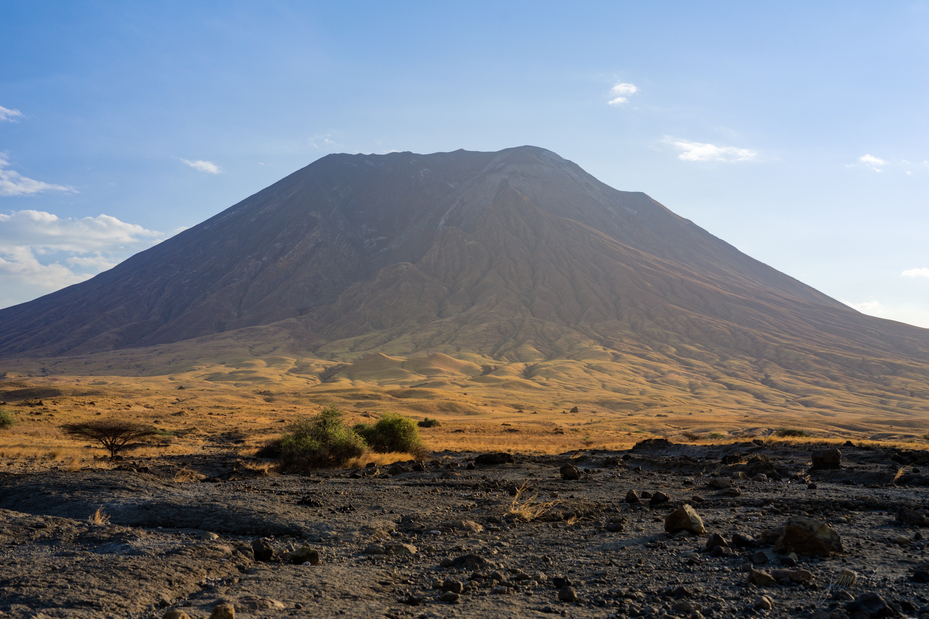 A steep grey volcanic cone rising from dry scrubland, its slopes streaked with pale ash, low afternoon light across the plain in front.