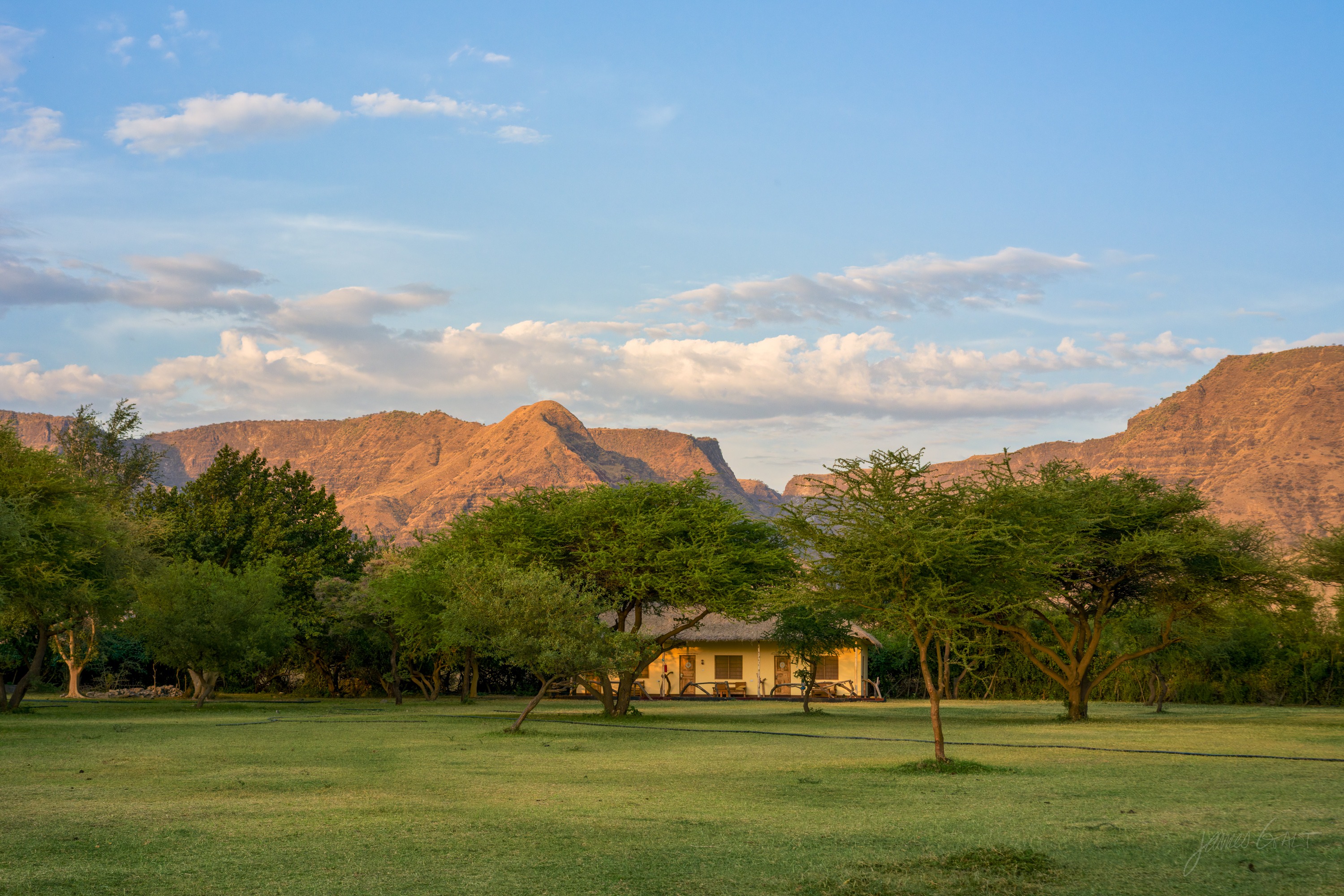 Lodge grounds in early golden light, a low building among trees and green lawn, long soft shadows across the ground.
