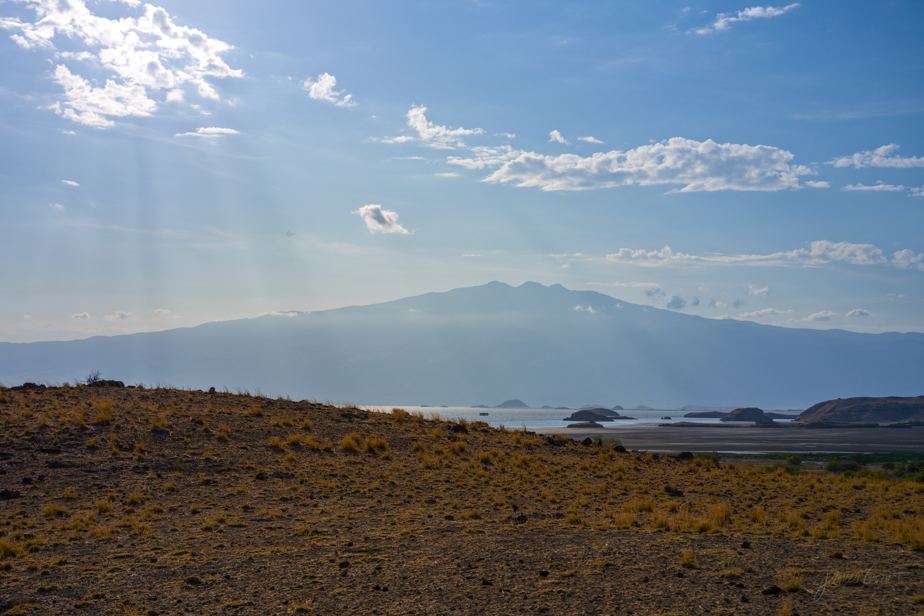 Sunlight breaking over the rim of a long escarpment, rays and haze in the air above a dry plain, the land below still in shadow.