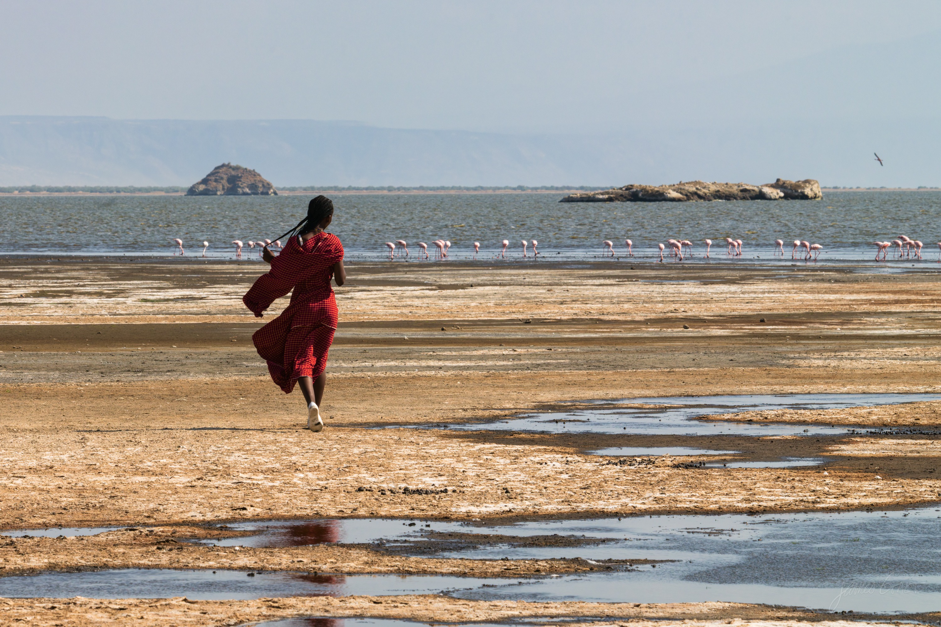 A person in a red robe walks alone across the pale cracked flats of the lake shore, a distant line of flamingos and a hazy shoreline beyond under a bright sky.