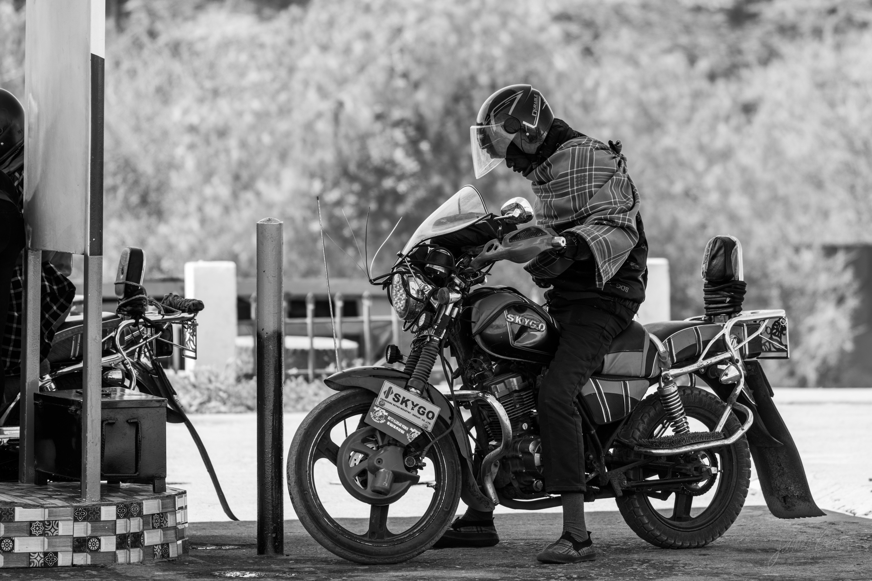 Black and white photograph of a person on a motorcycle pulled up at a small roadside fuel station, luggage strapped behind them, pumps and painted posts framing the scene.