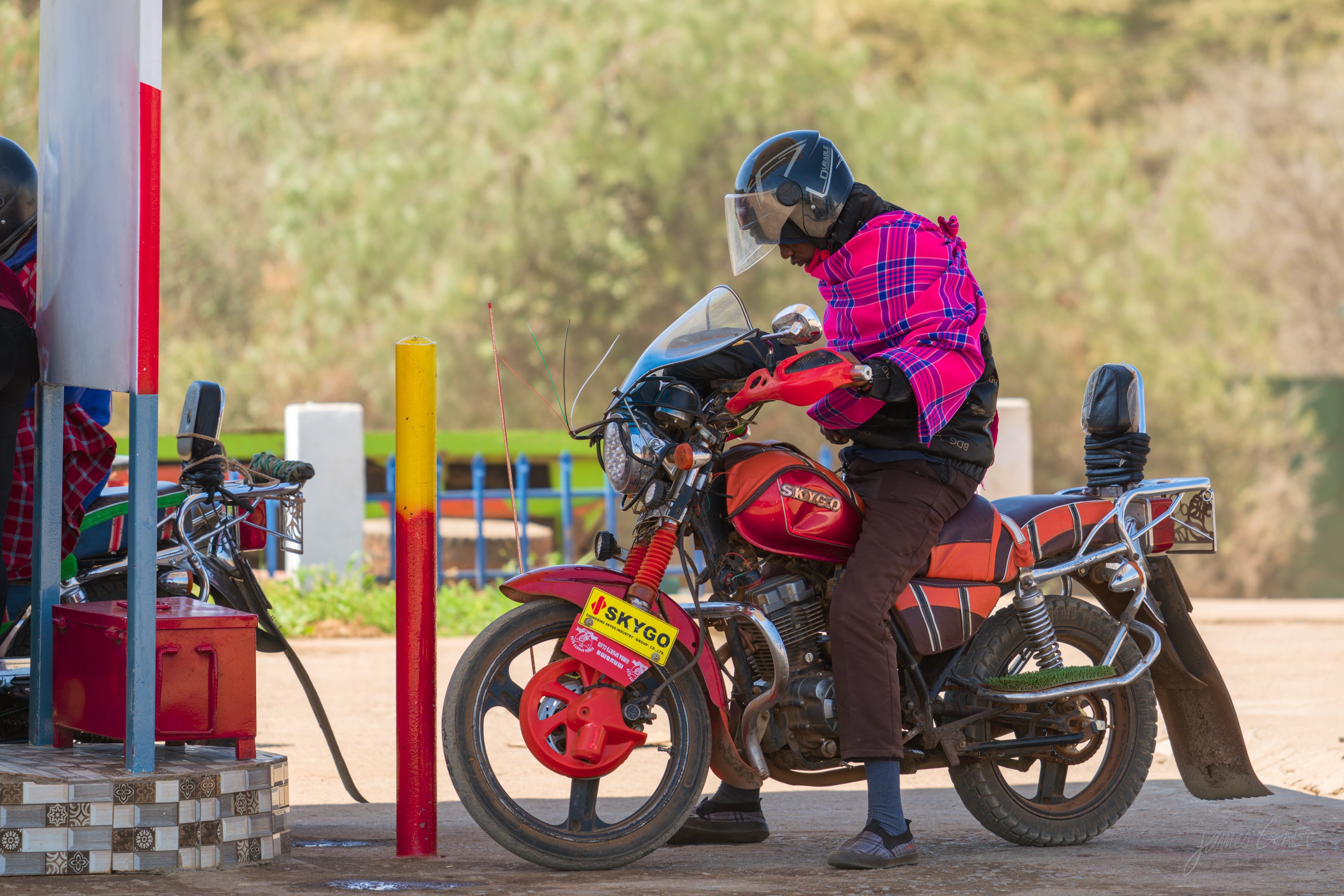 The same scene in colour, a rider in a pink checked shirt on a red motorcycle at a fuel station with red and yellow pumps and brightly painted posts.