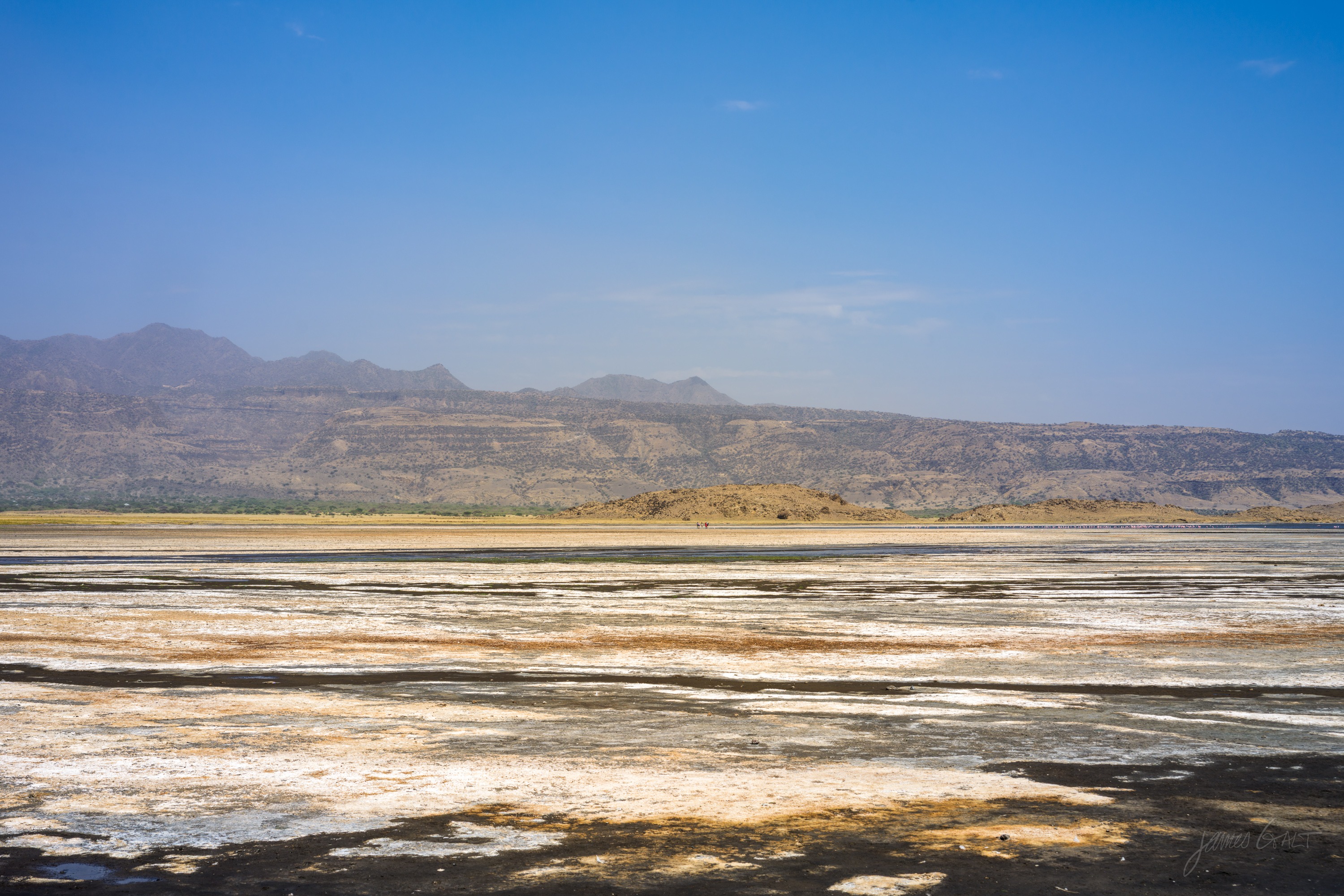 A wide flat expanse of pale salt and mud stretching to a low line of hills, thin sheets of water catching light across the middle distance.