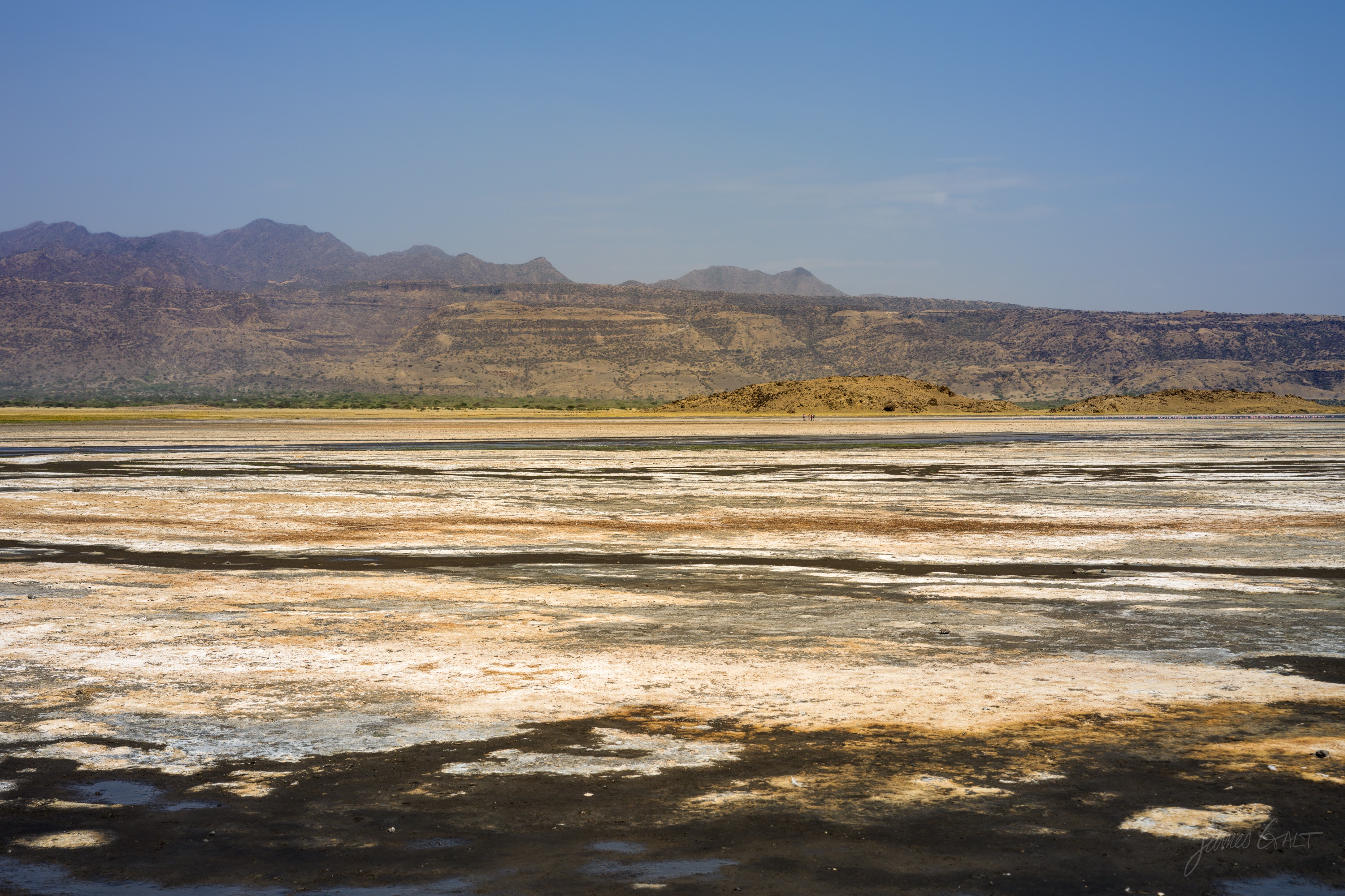 The shore of the soda lake, pale crusted flats in the foreground giving way to shallow water and a hazy far bank under a bright sky.