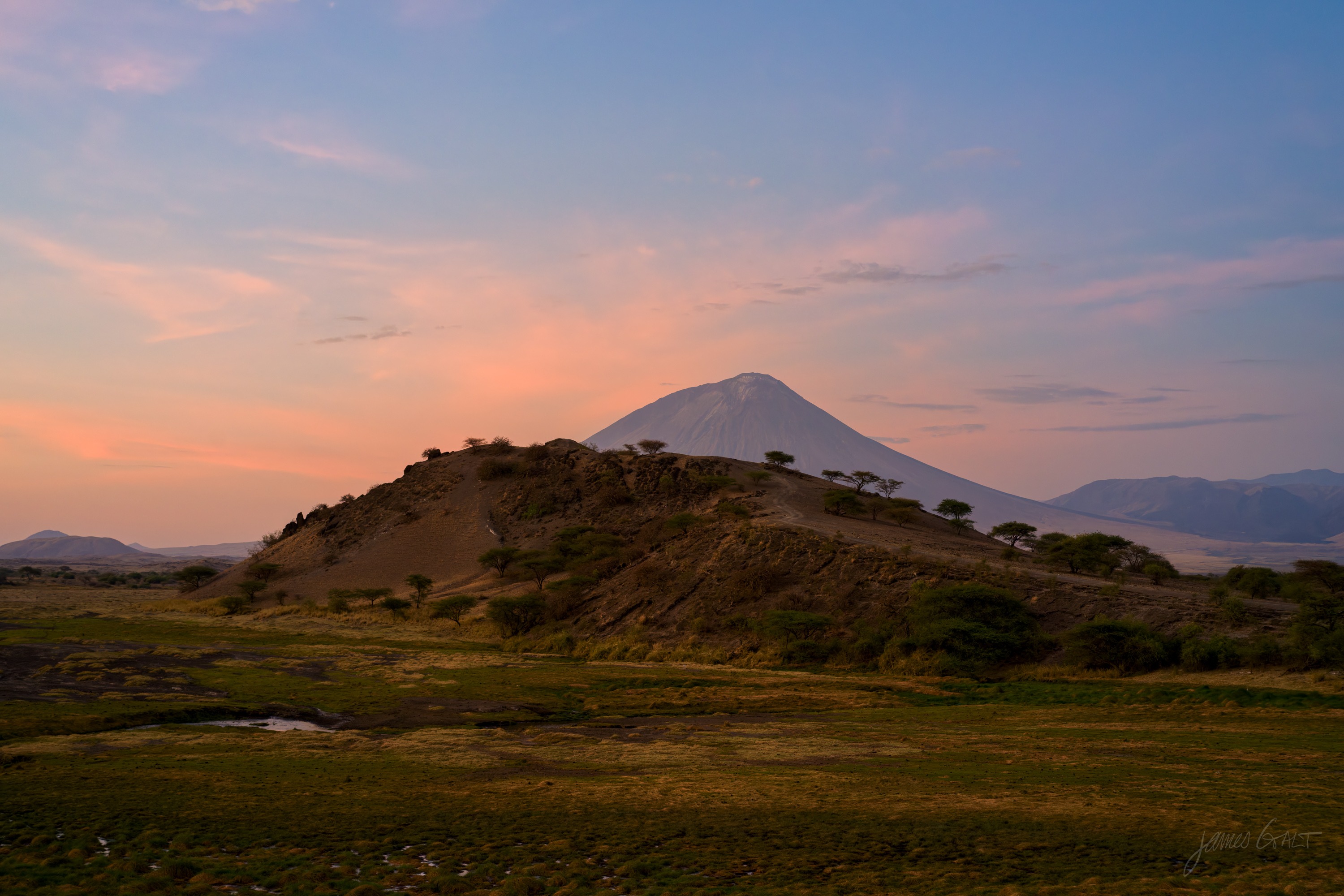 A dark volcanic cone silhouetted against a soft orange and pink dawn sky, the plain below still in shadow.