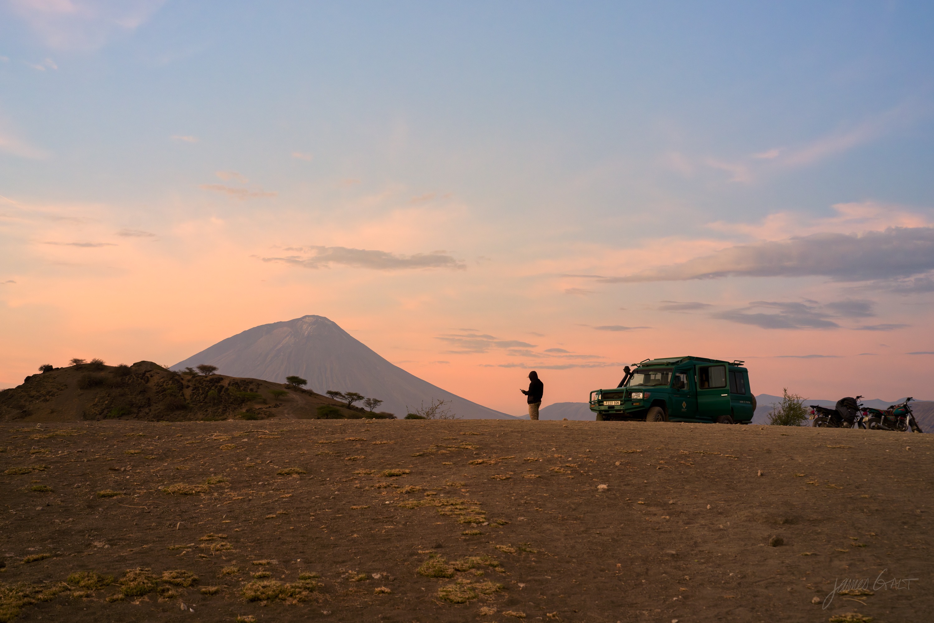 A safari vehicle parked on the plain at dawn with a small figure standing beside it, both silhouetted against a volcanic cone and a glowing sky.