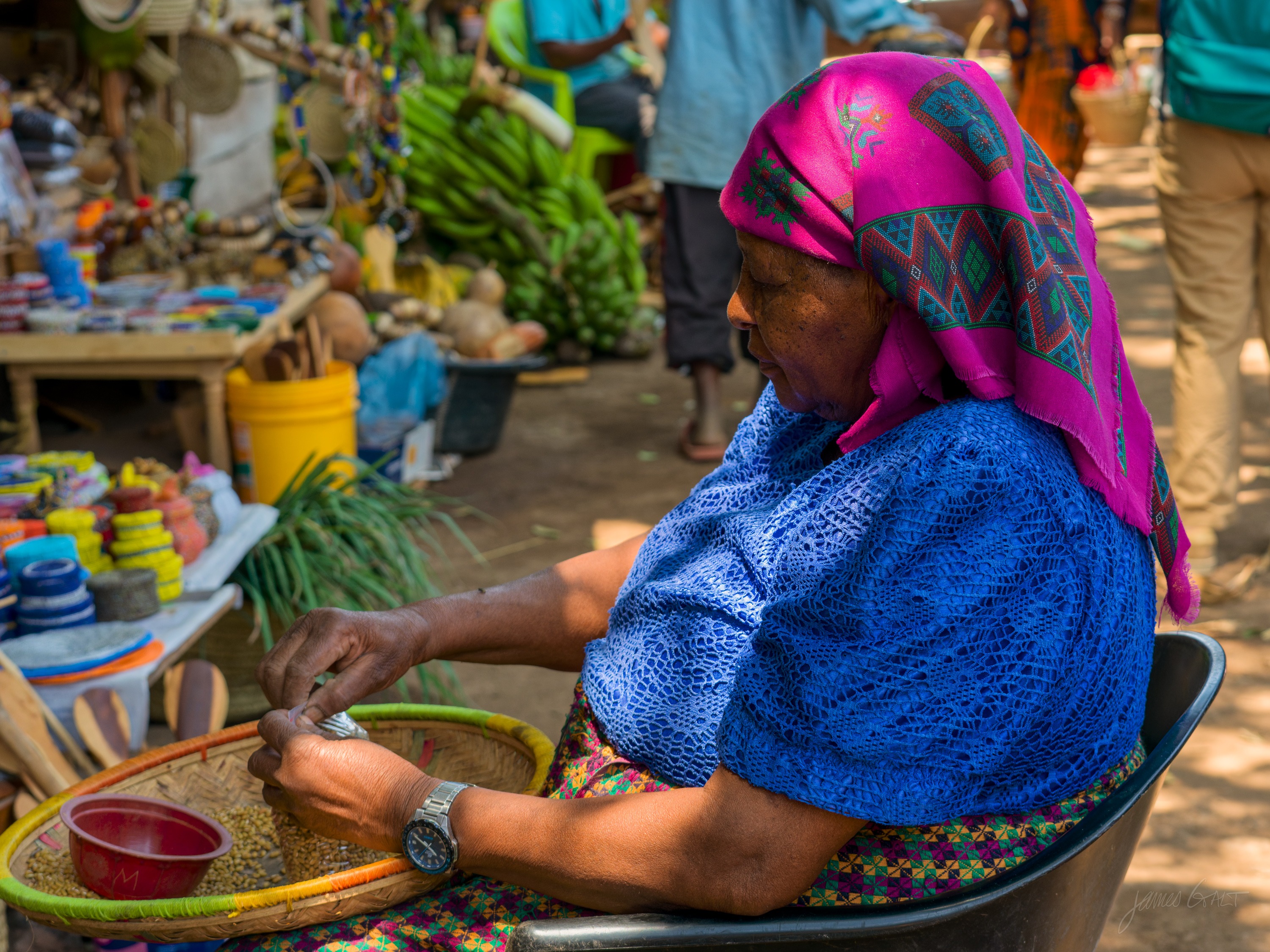 A woman in a bright pink headscarf and blue top sits weaving a coiled basket, a shallow bowl and lengths of coloured fibre in front of her, a stall of goods blurred behind.