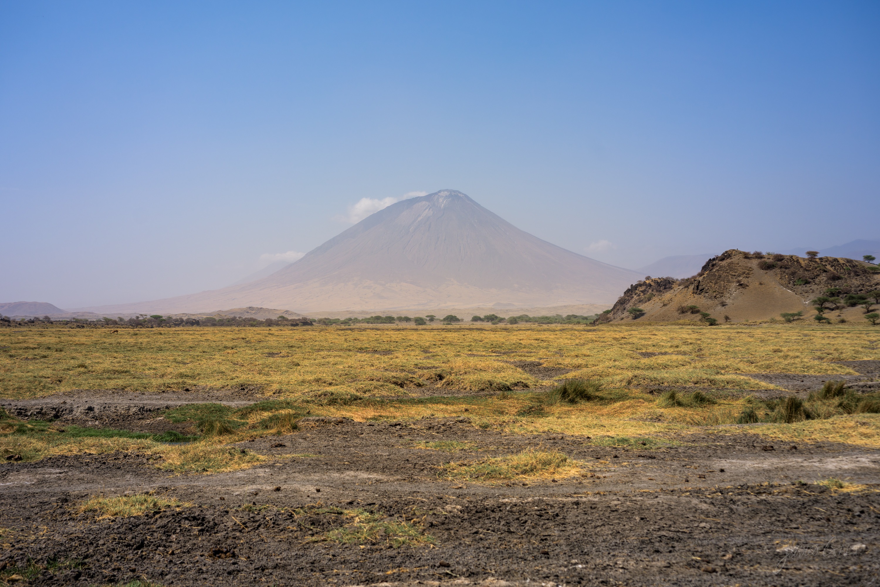 A grey volcanic cone rising beyond a wide green and gold plain, low scrub across the foreground and a hazy sky, flat midday light on the land.
