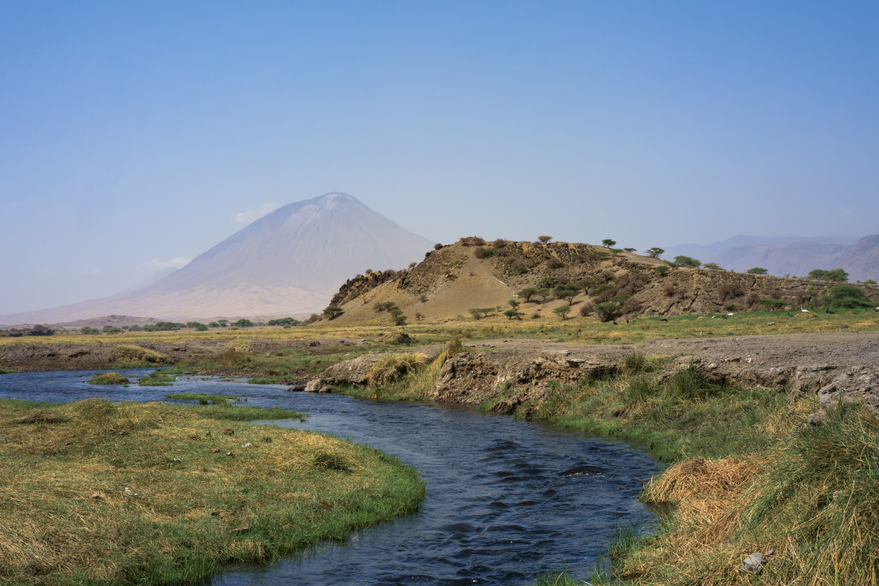 A narrow green stream curves through dry plain toward the base of a steep volcanic cone, reeds along its banks, the mountain pale in the haze behind.