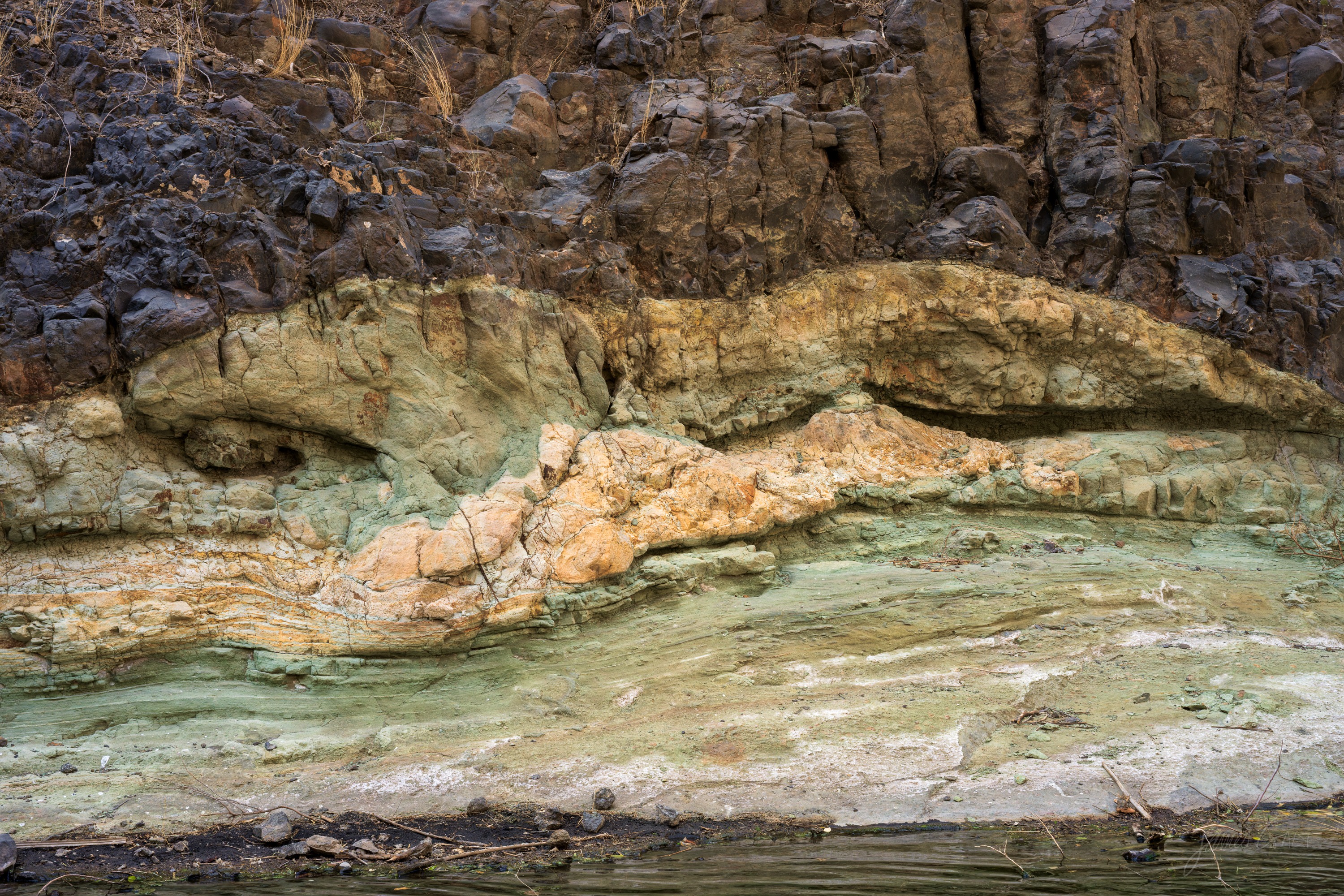 A close view of layered, water-worn rock in warm ochre and green tones, the bands folding across the face of a small gorge.