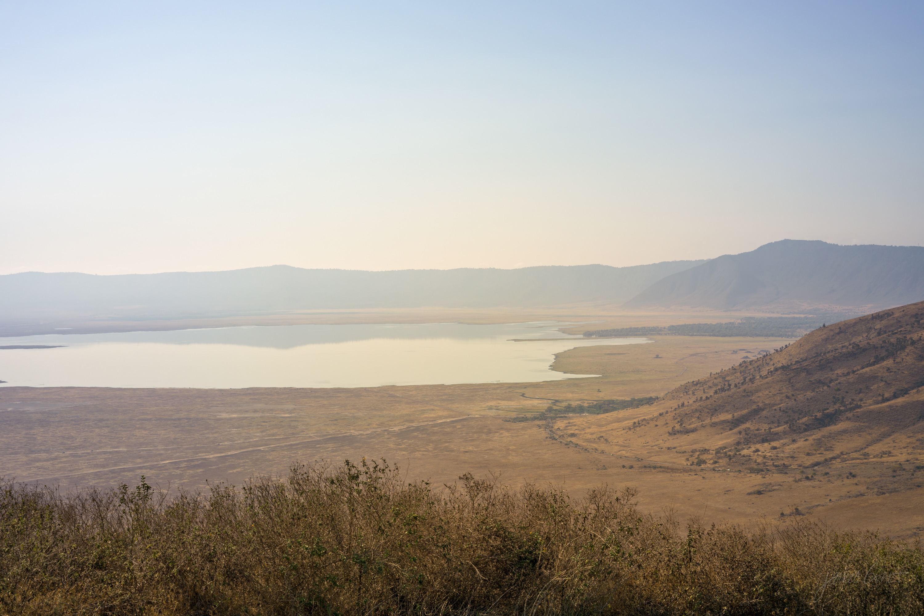A wide view over the Ngorongoro crater floor from the rim, a patchwork of green and gold plain with a pale lake in the distance under a high sky.