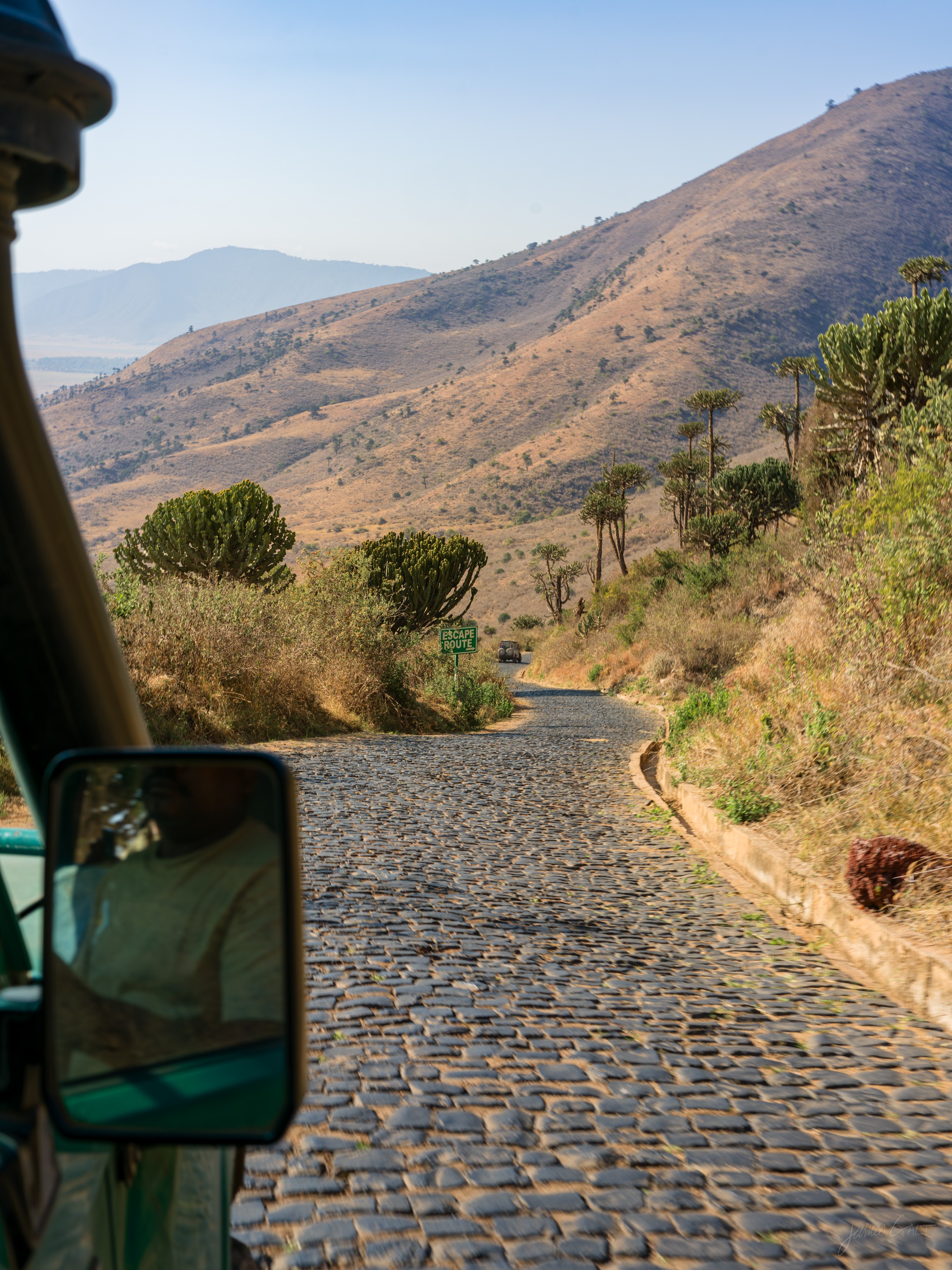 A narrow cobbled road descending steeply into the crater, hills rising alongside, the wing mirror of a safari vehicle in the corner of the frame.