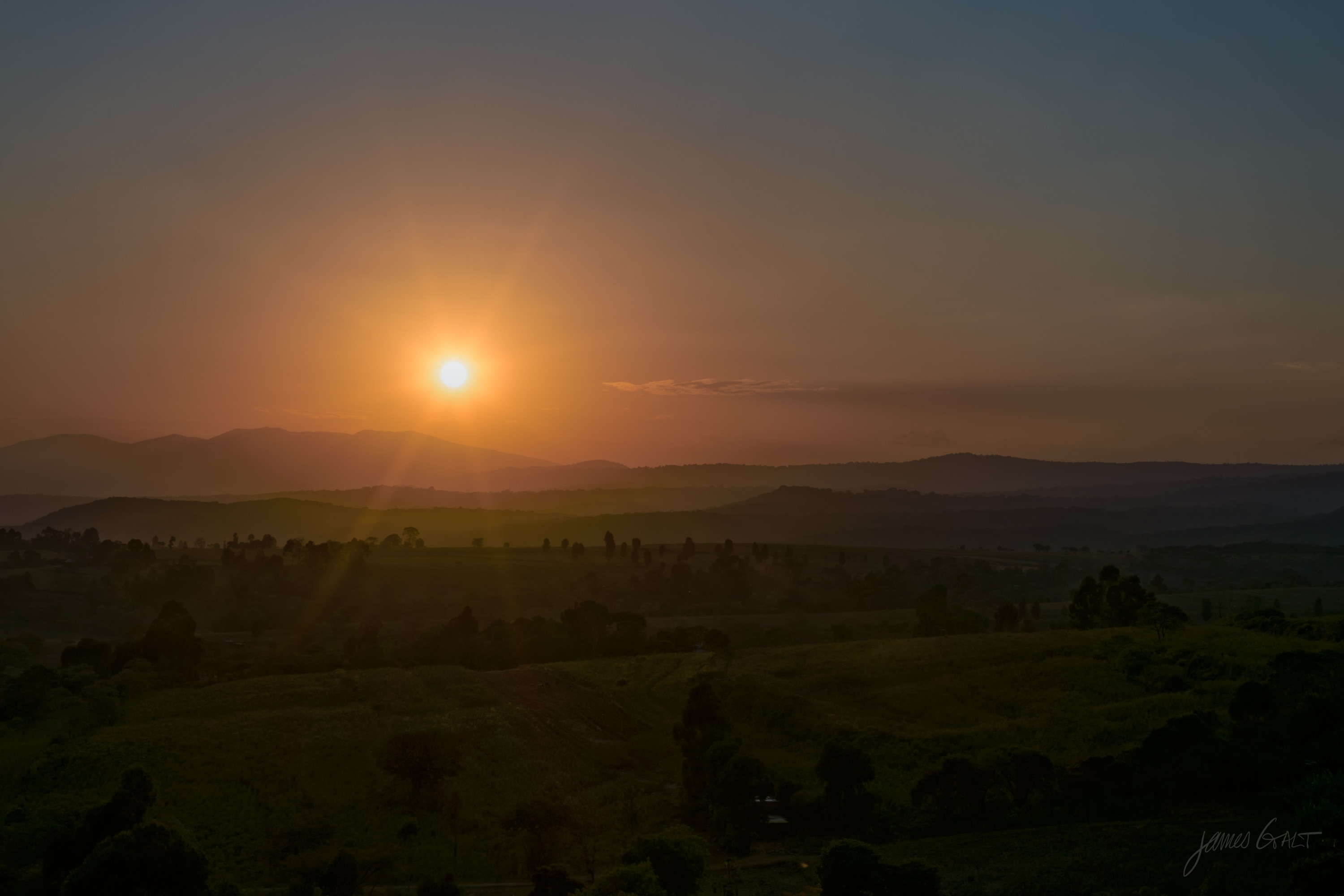 Low hills and scattered trees going blue and gold in the last light of the day, the sun low behind a hazy horizon.