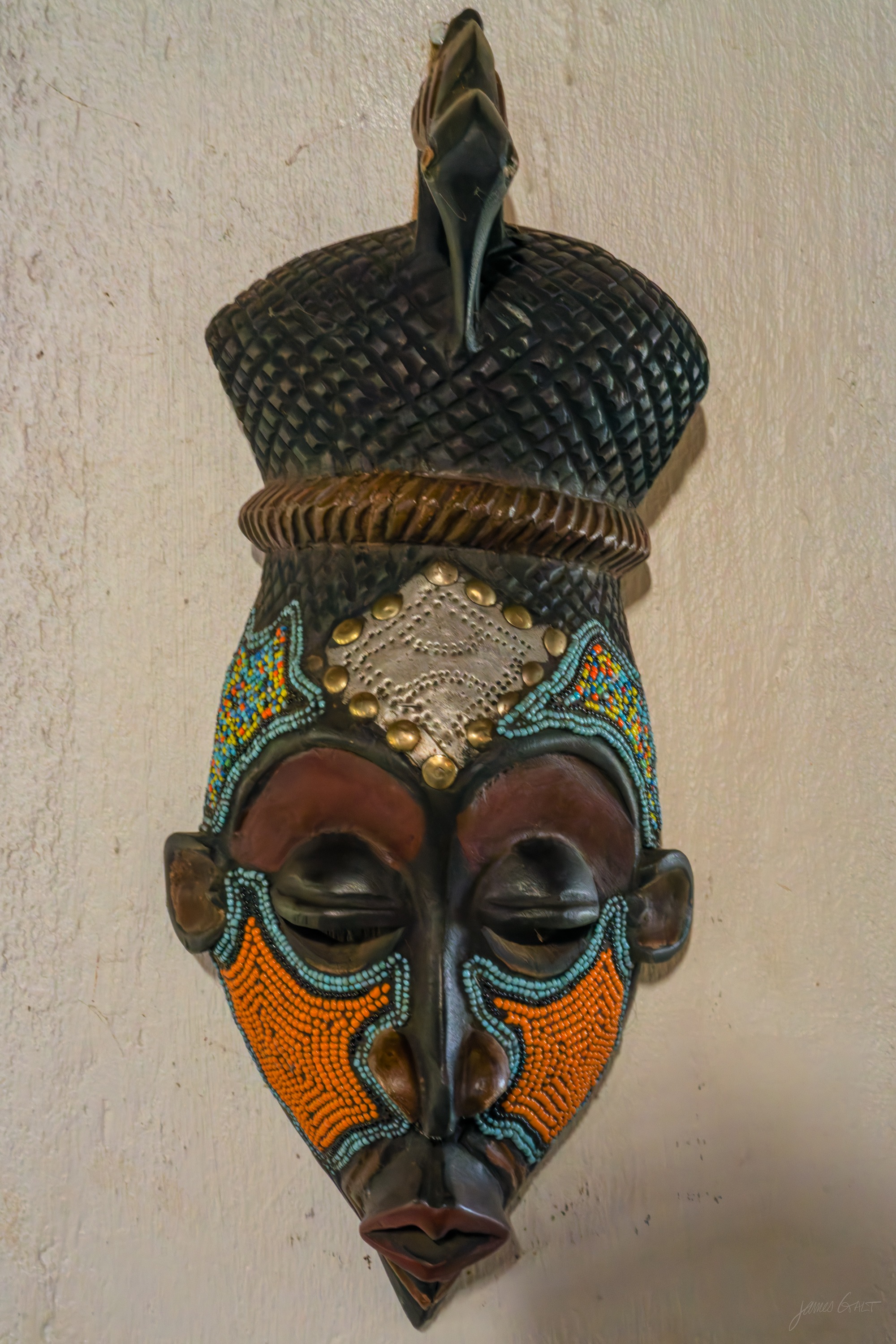 A carved mask entirely covered in tiny coloured beads forming patterns across an elongated face, mounted on a pale wall in dim light.