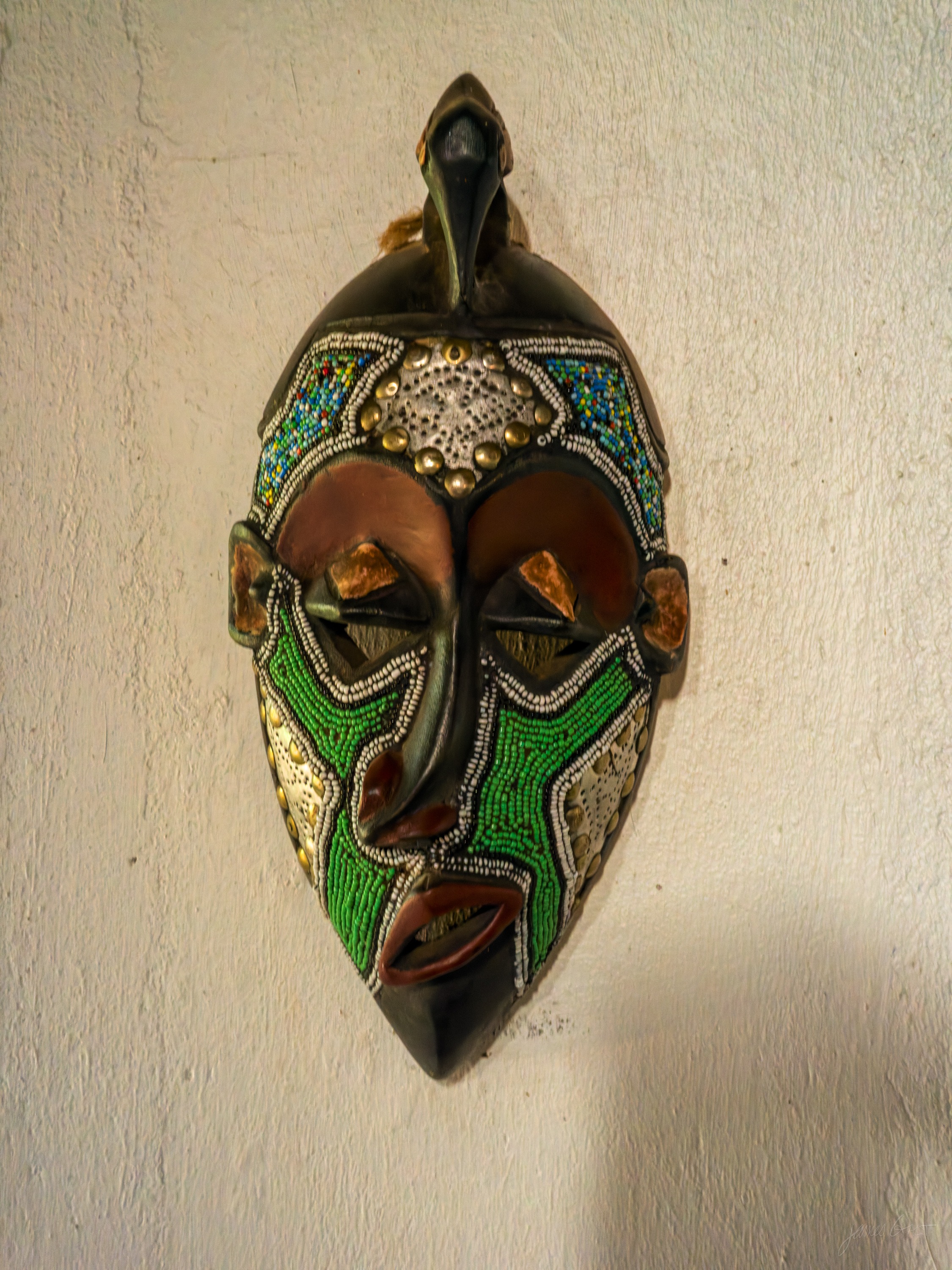 A second beaded mask with a broad face worked in green, orange and dark beads, the surface catching a little light in the gloom.