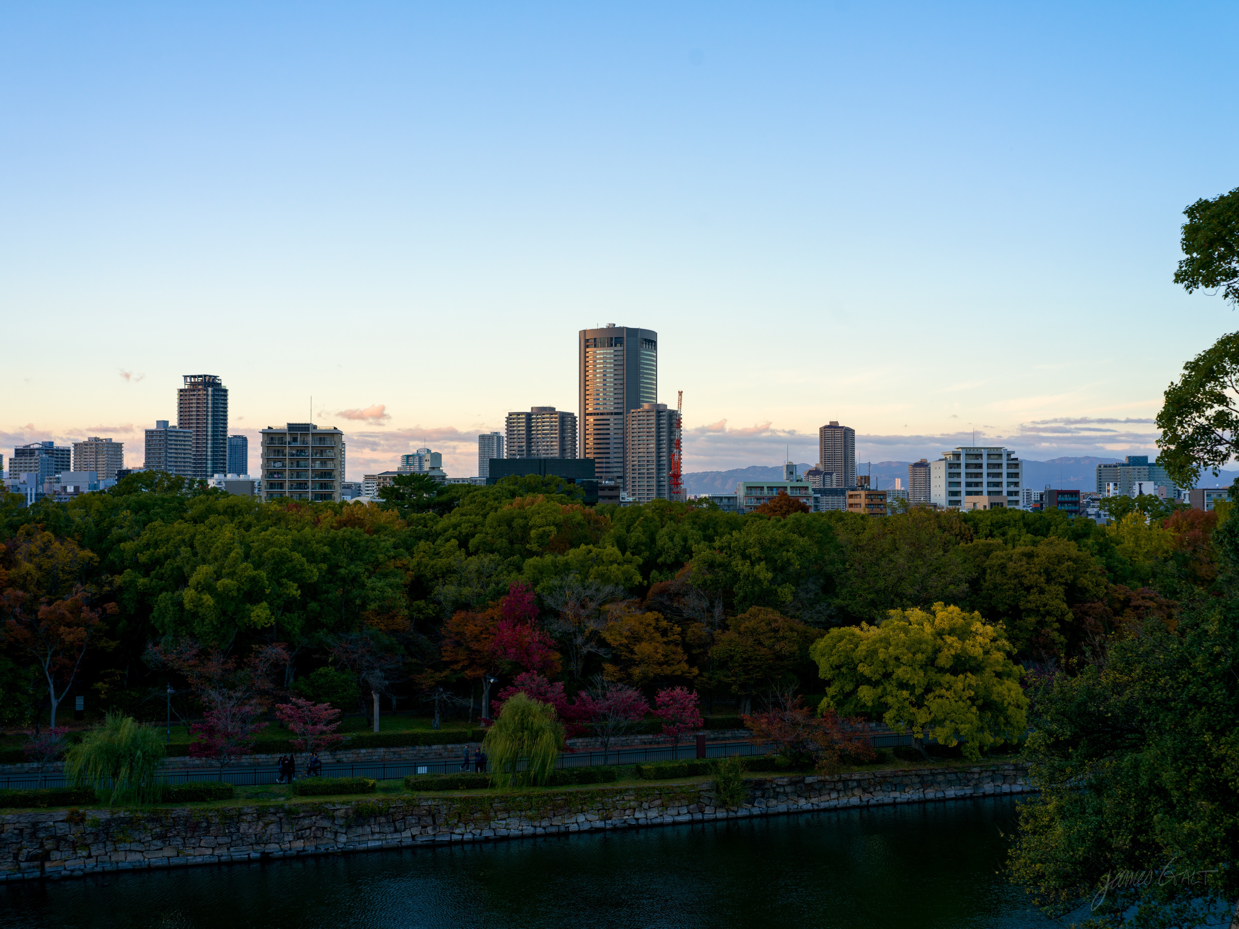 A late-afternoon view across the autumn castle park, trees turning red, orange and yellow above a stone-walled moat with willows. Apartment and office towers and distant mountains sit on the horizon under a pale blue sky.