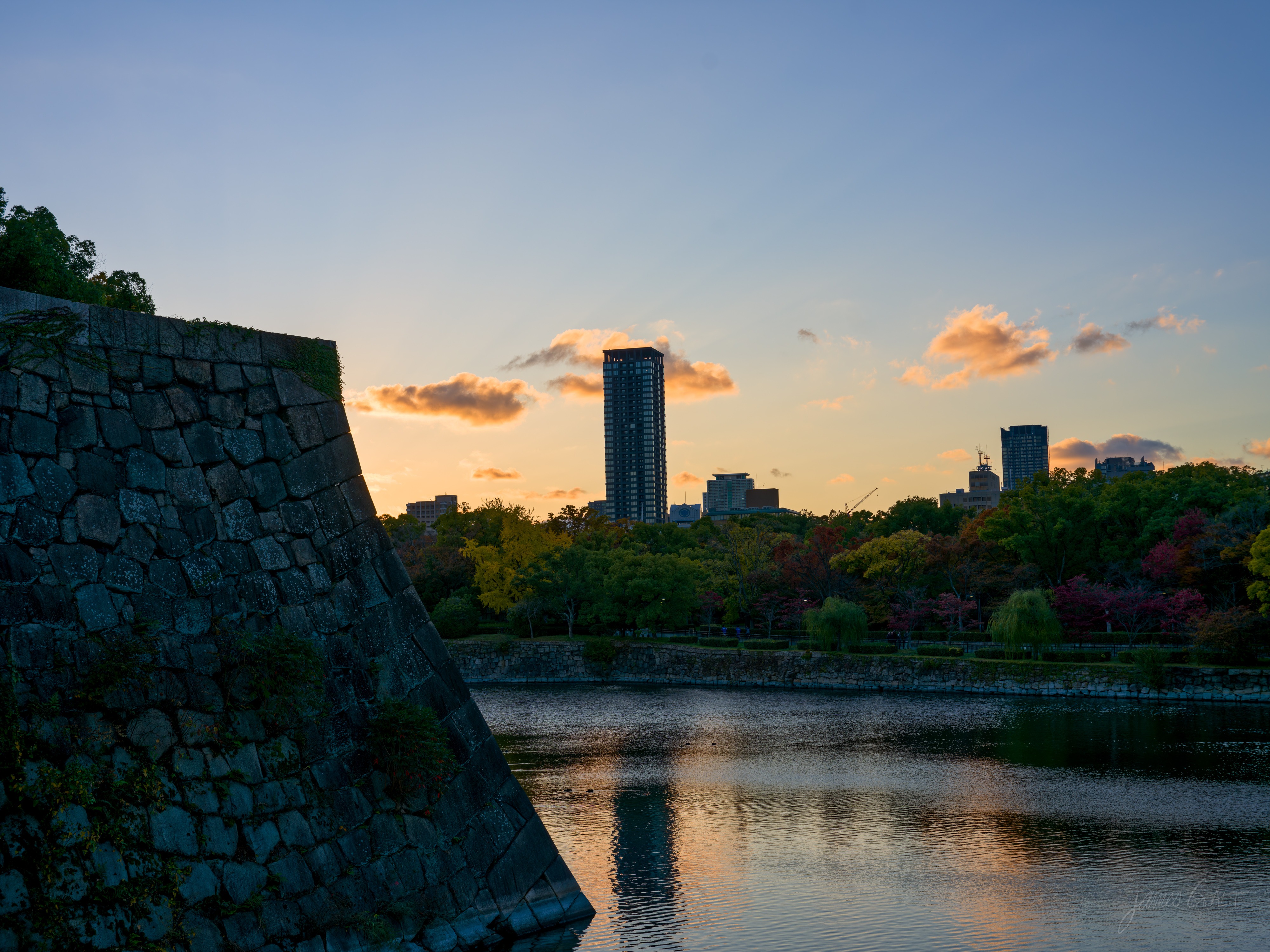 Golden hour at the castle moat: a huge sloping stone rampart in shadow on the left, calm water reflecting an orange sky, and a tall apartment tower lit by the setting sun above autumn trees on the far bank.