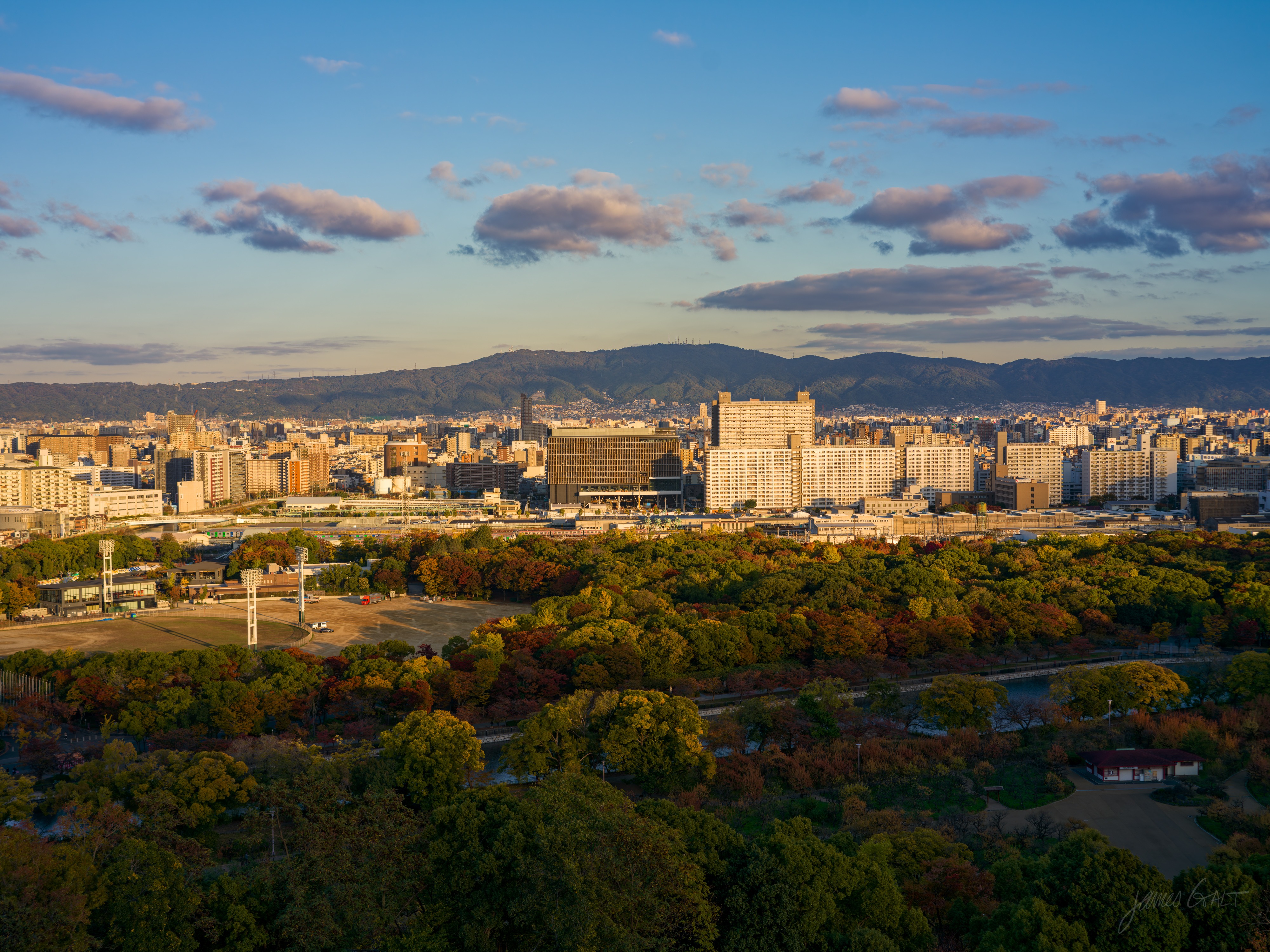 A high view over the green castle park, its trees turning autumn colours, toward a long pale institutional building and the dense low city beyond, with a range of mountains on the horizon under a blue sky streaked with cloud, all warm in the late sun.