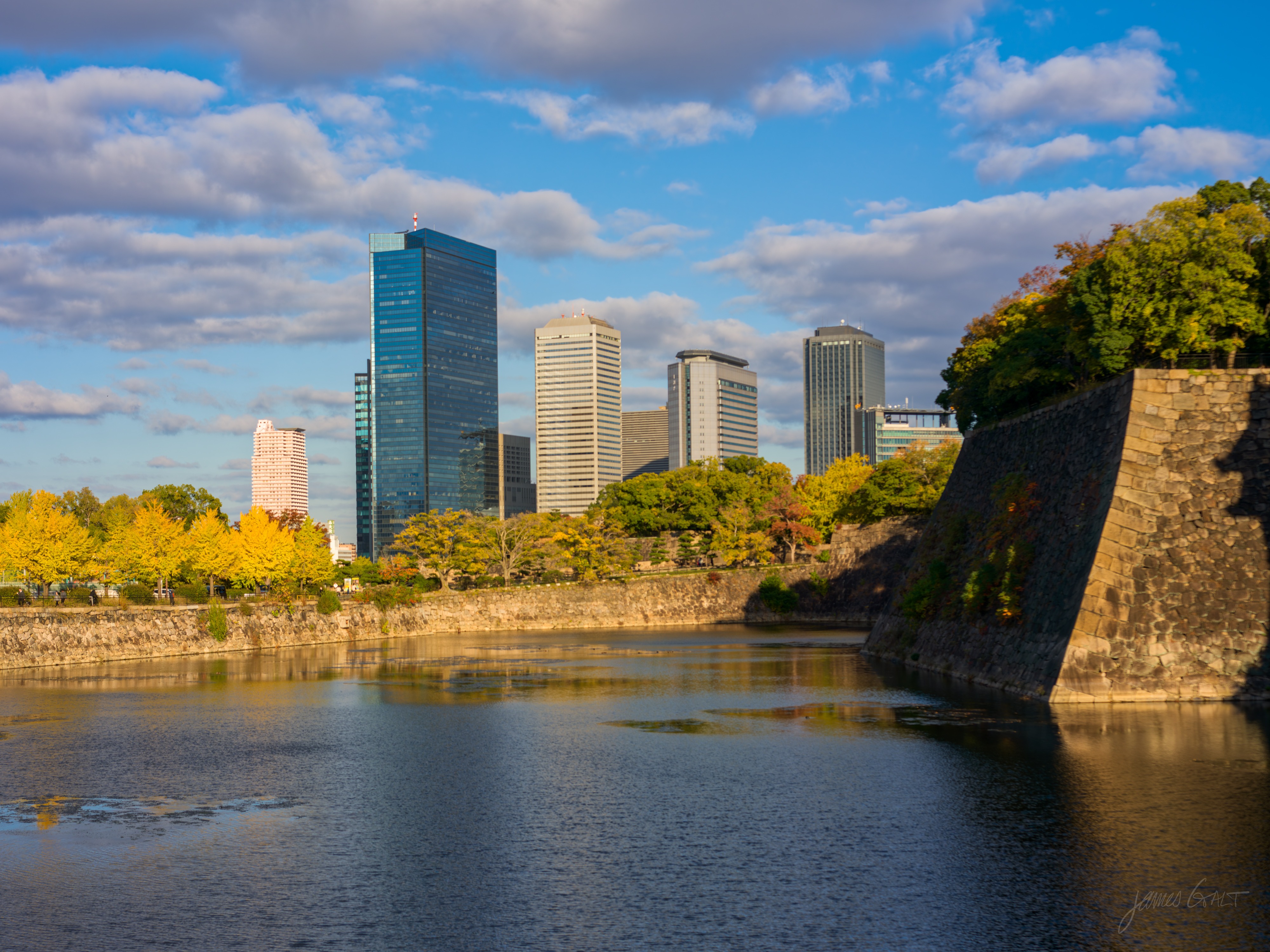 The moat of Osaka Castle in afternoon light, a massive sloping stone rampart on the right and a low stone wall with yellow autumn trees on the left. Behind them rise the glass office towers of the Business Park under a blue sky with scattered cloud, reflected in the calm water.