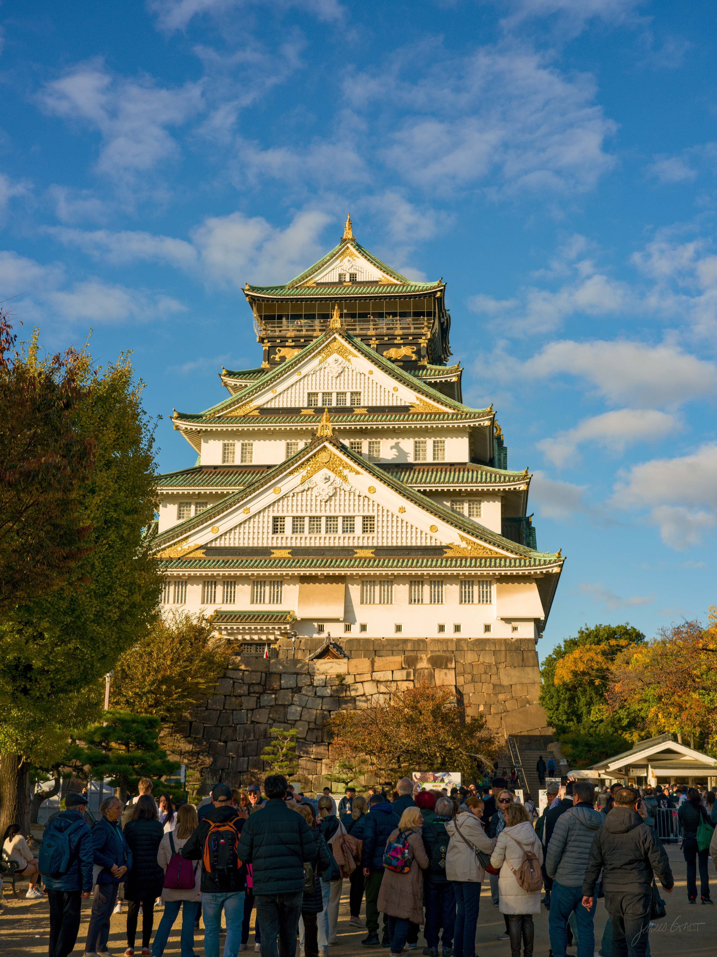The main keep of Osaka Castle lit by low afternoon sun, its white walls, green-tiled roofs and gold trim rising above a stone base, framed by autumn trees. A large crowd of visitors gathers on the plaza in front.