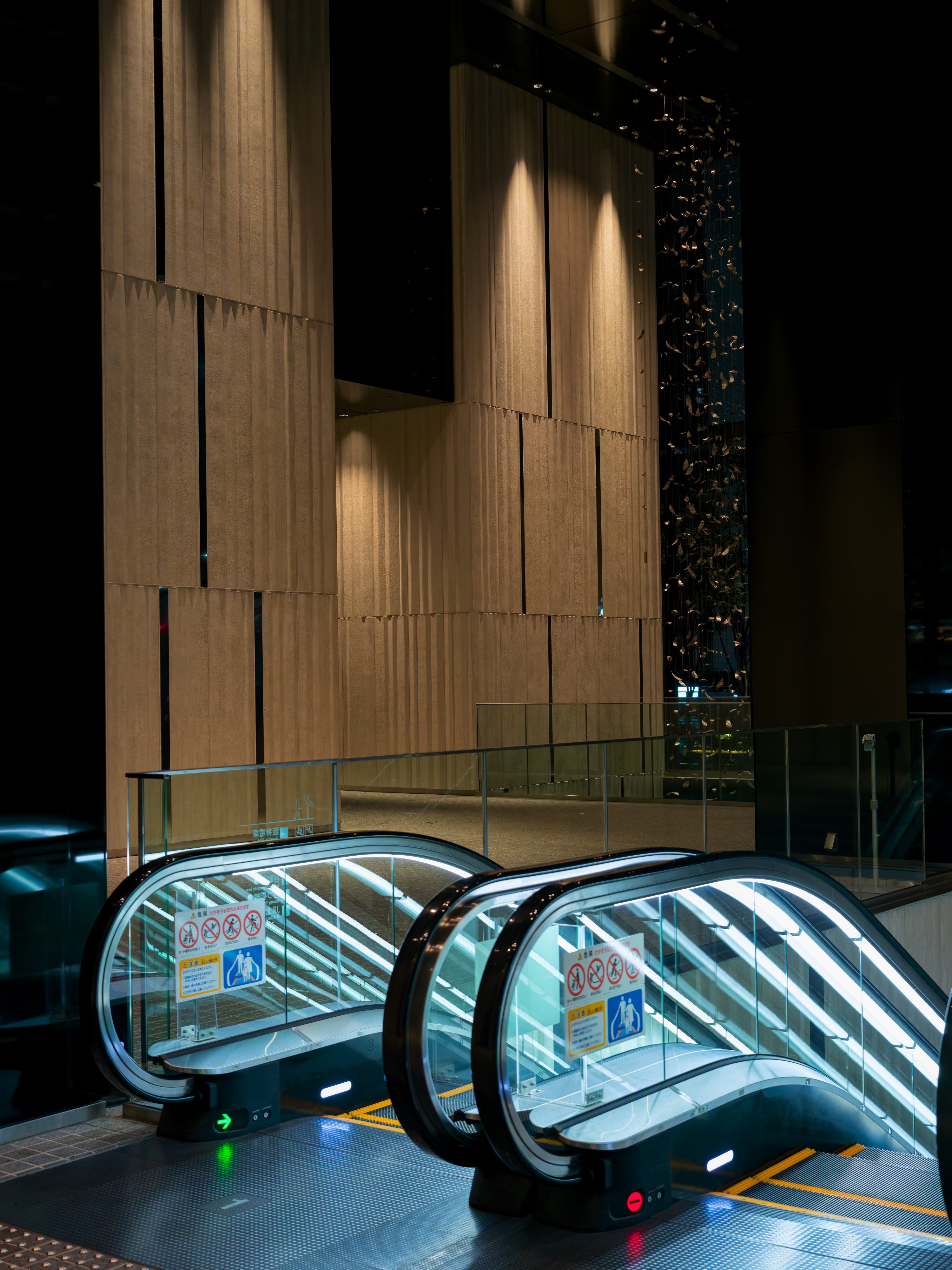 Two parallel escalators with glass balustrades lit from within, rising toward a tall beige ribbed wall at night, dark glass beside them showing distant city lights. Directional arrows mark the floor.