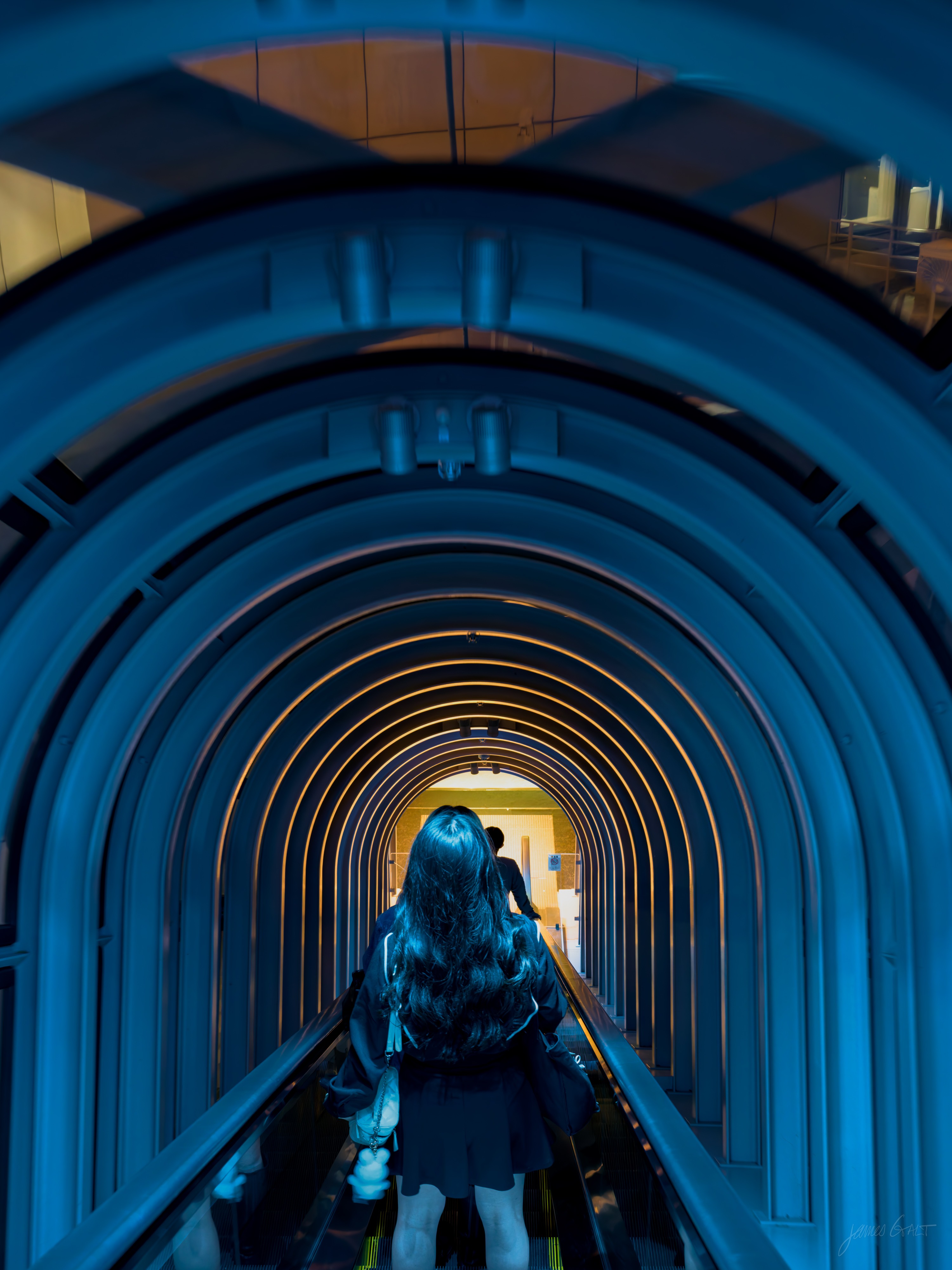 Inside a diagonal glass tube escalator at night, the arched structural ribs lit blue and receding to a small golden opening far ahead. A woman with long hair, seen from behind, rides up toward the light, another figure ahead of her.