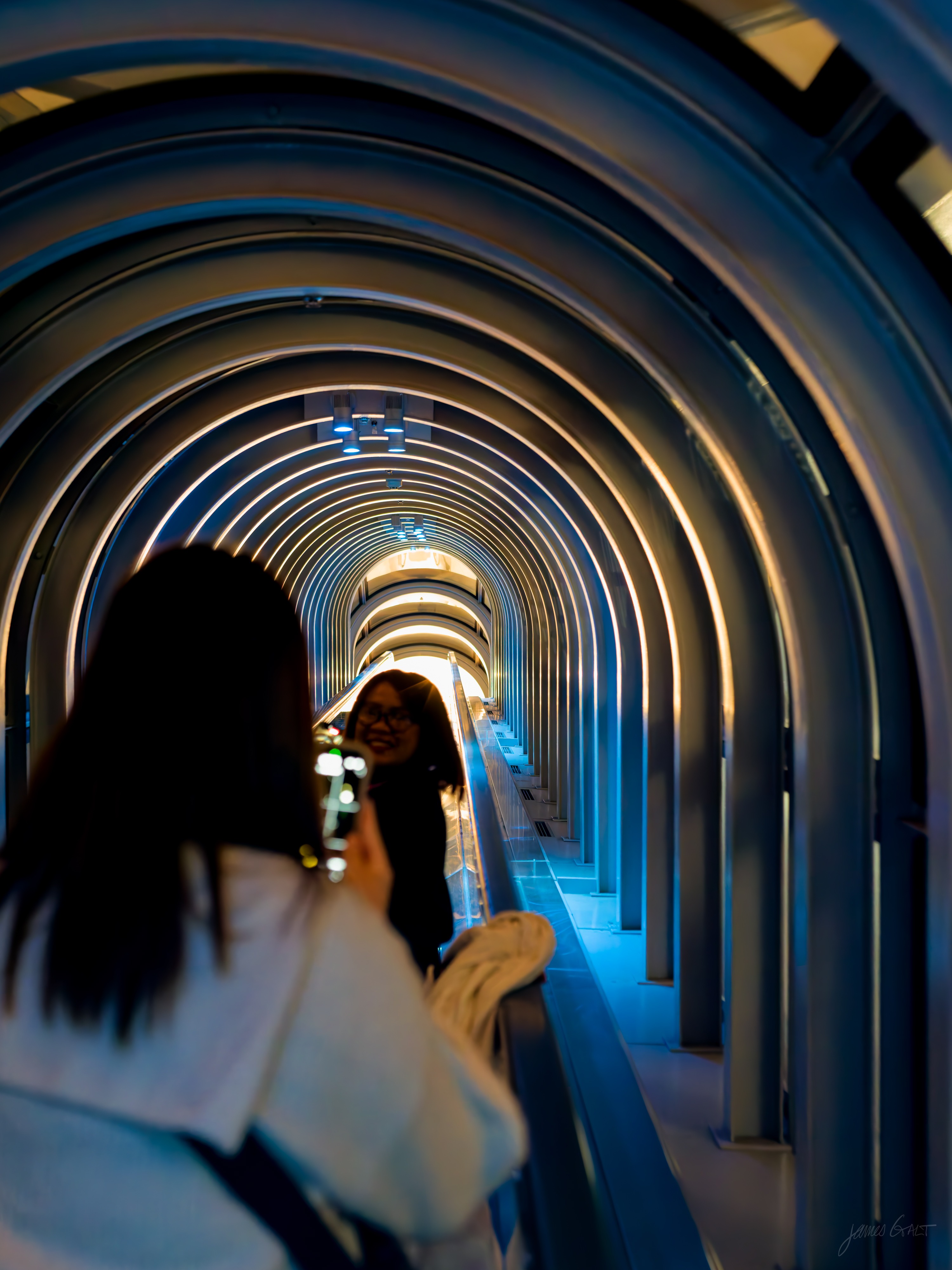 Inside the same blue-lit escalator tube, a woman in the foreground (out of focus, back to camera) holds up a phone to photograph a second woman, who leans on the handrail further along and smiles back, the arched ribs curving around them.