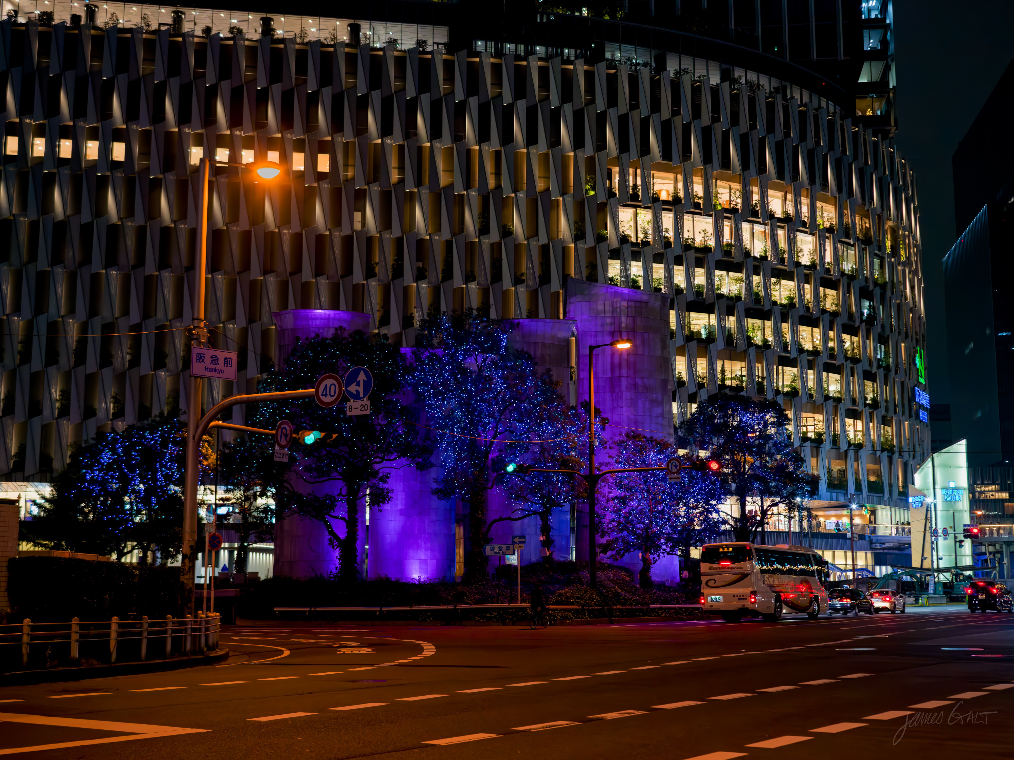 A wide, near-empty city intersection at night, a large curved building rising behind it. At street level, trees and low pillars are lit purple and blue, lit office windows stacked above, with a few cars and a bus on the road.