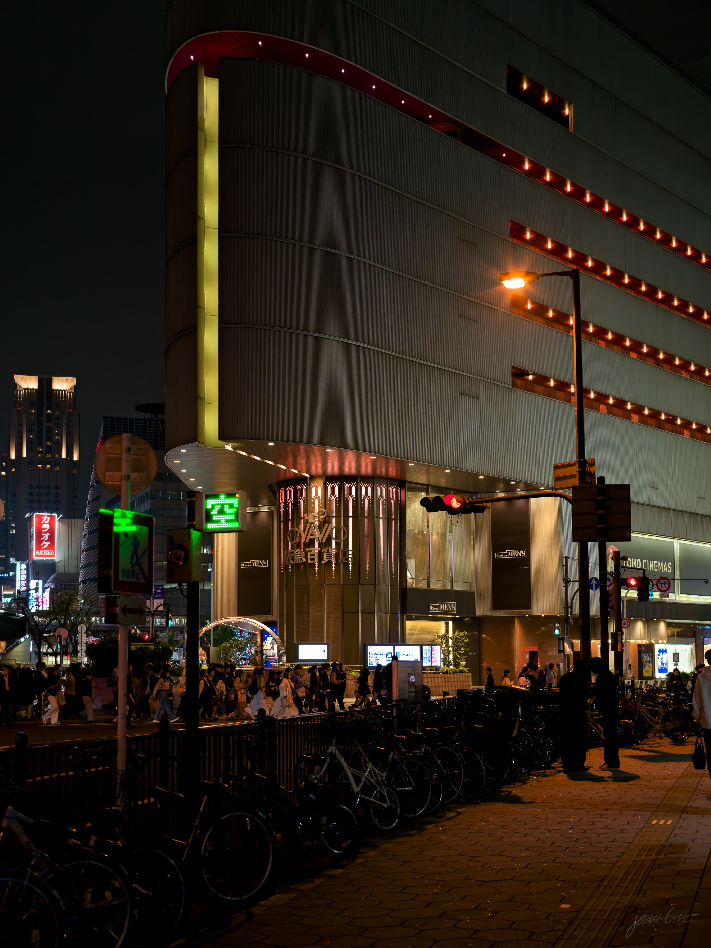 A night street beside a large department-store building with red string lights tracing its curved upper floors and a yellow vertical strip down the corner. Green pedestrian signals glow, a long row of bicycles is parked along a railing, and a crowd gathers near the entrance and a cinema.