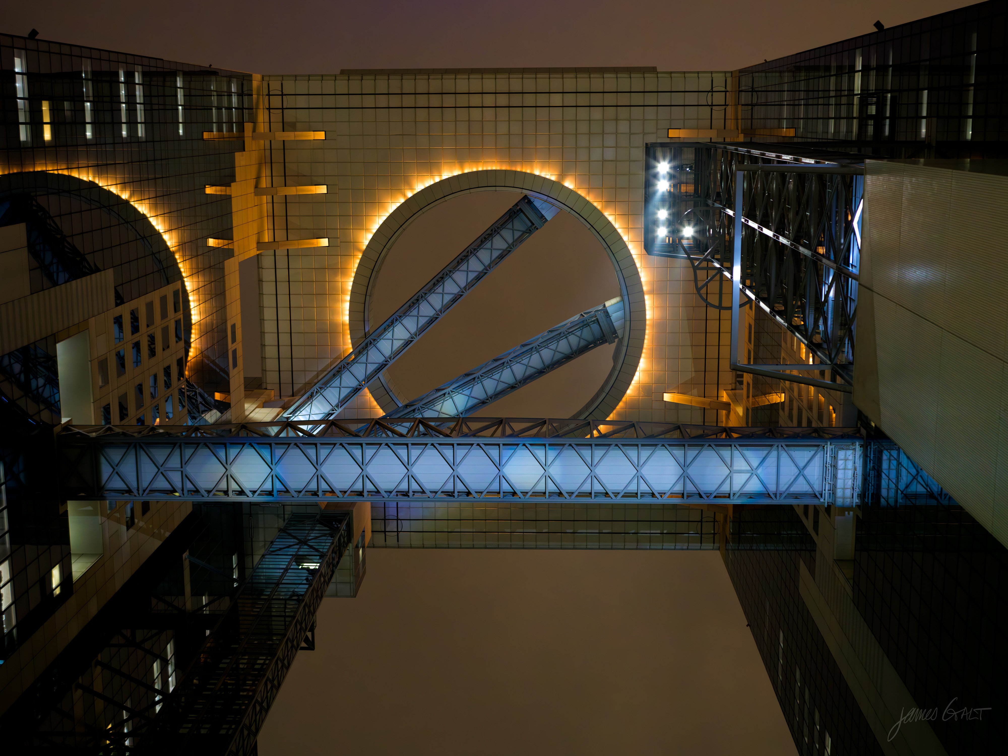 Looking straight up through the open gap between the two towers of the Umeda Sky Building at night. A large ring-shaped rooftop observatory is lit gold, with diagonal escalators and a blue-lit glass sky bridge crossing the void below it, framed by the dark glass walls of the towers.