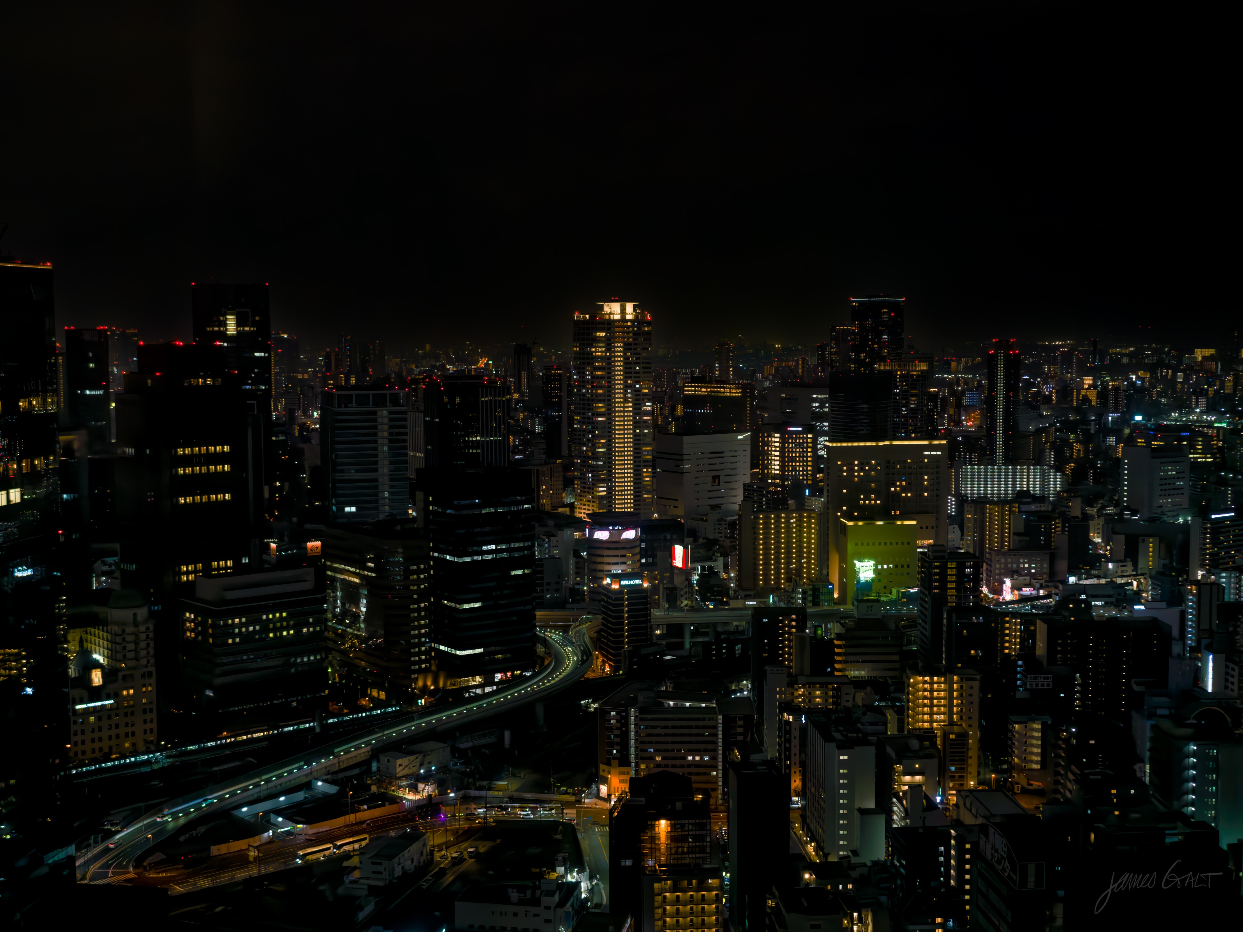 A night view over Osaka from high up: a dense field of low buildings and scattered lit towers under a black sky, with a curving elevated expressway picked out in light threading through the middle.