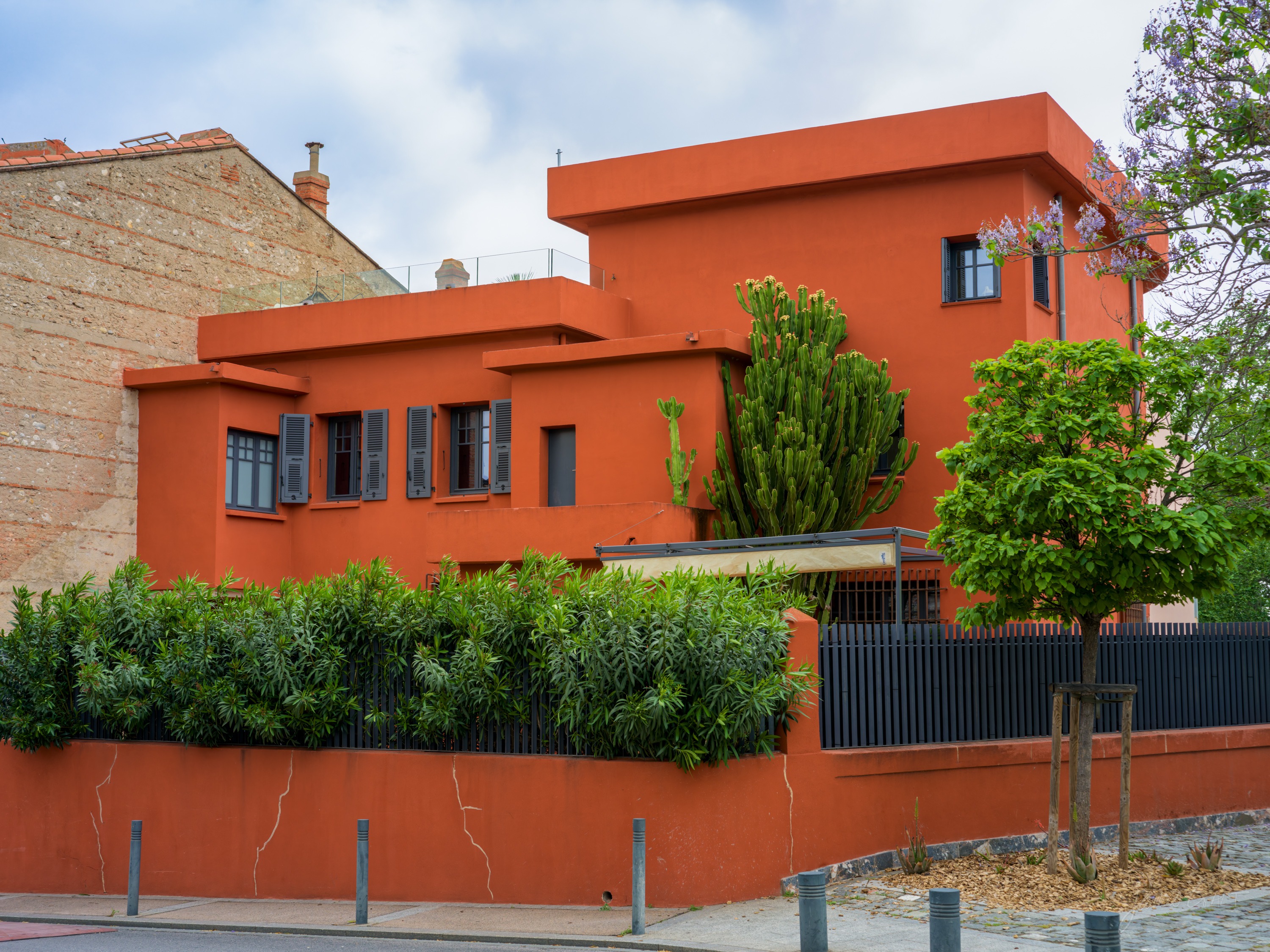 A bold orange-red modernist villa with flat roofs and grey shutters, a tall cactus and clipped greenery over its garden wall, a cracked render wall to the left.