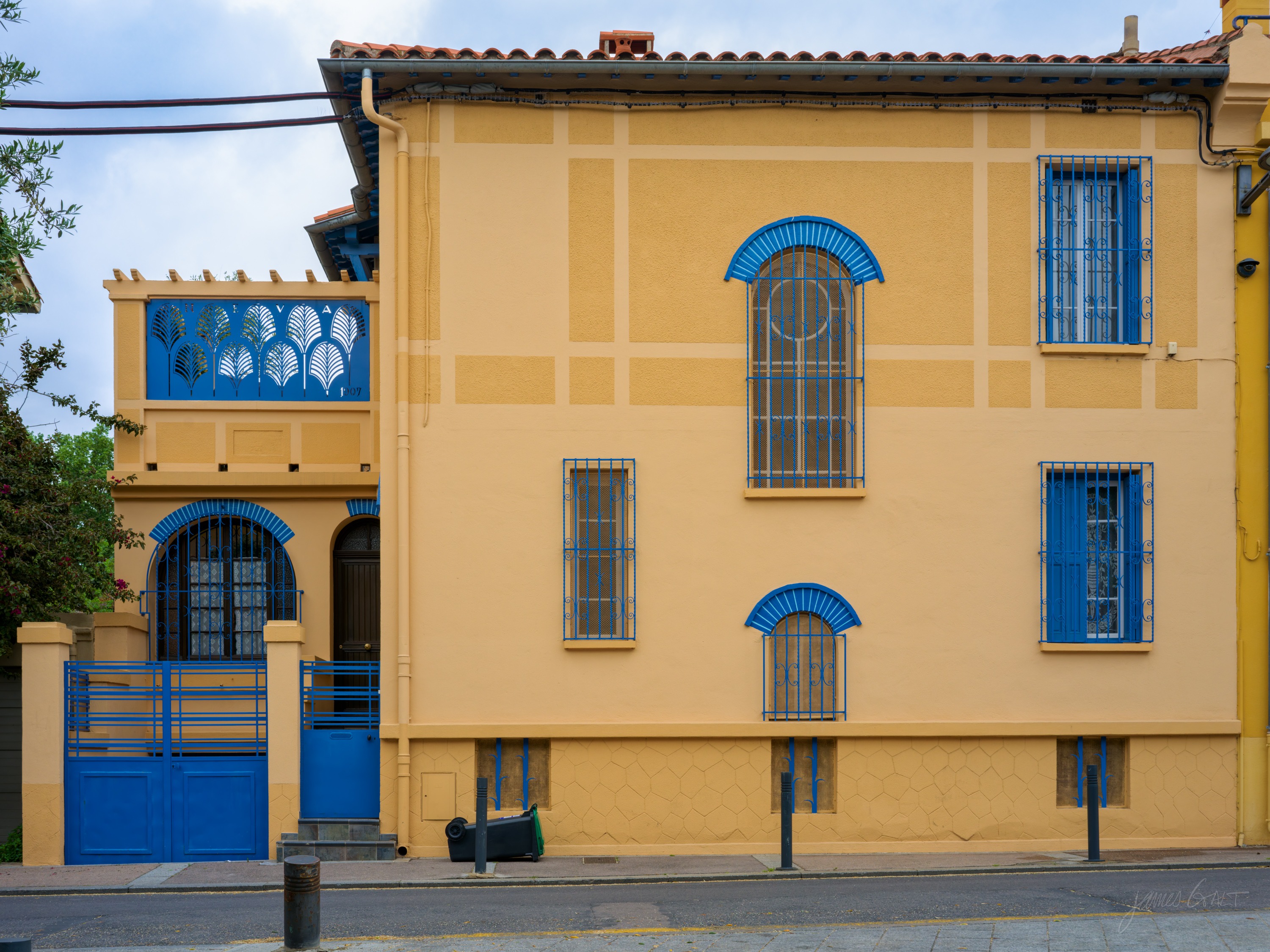 A yellow ochre house with Art Nouveau windows outlined in bright blue, a matching blue gate and a small tiled loggia at one corner.