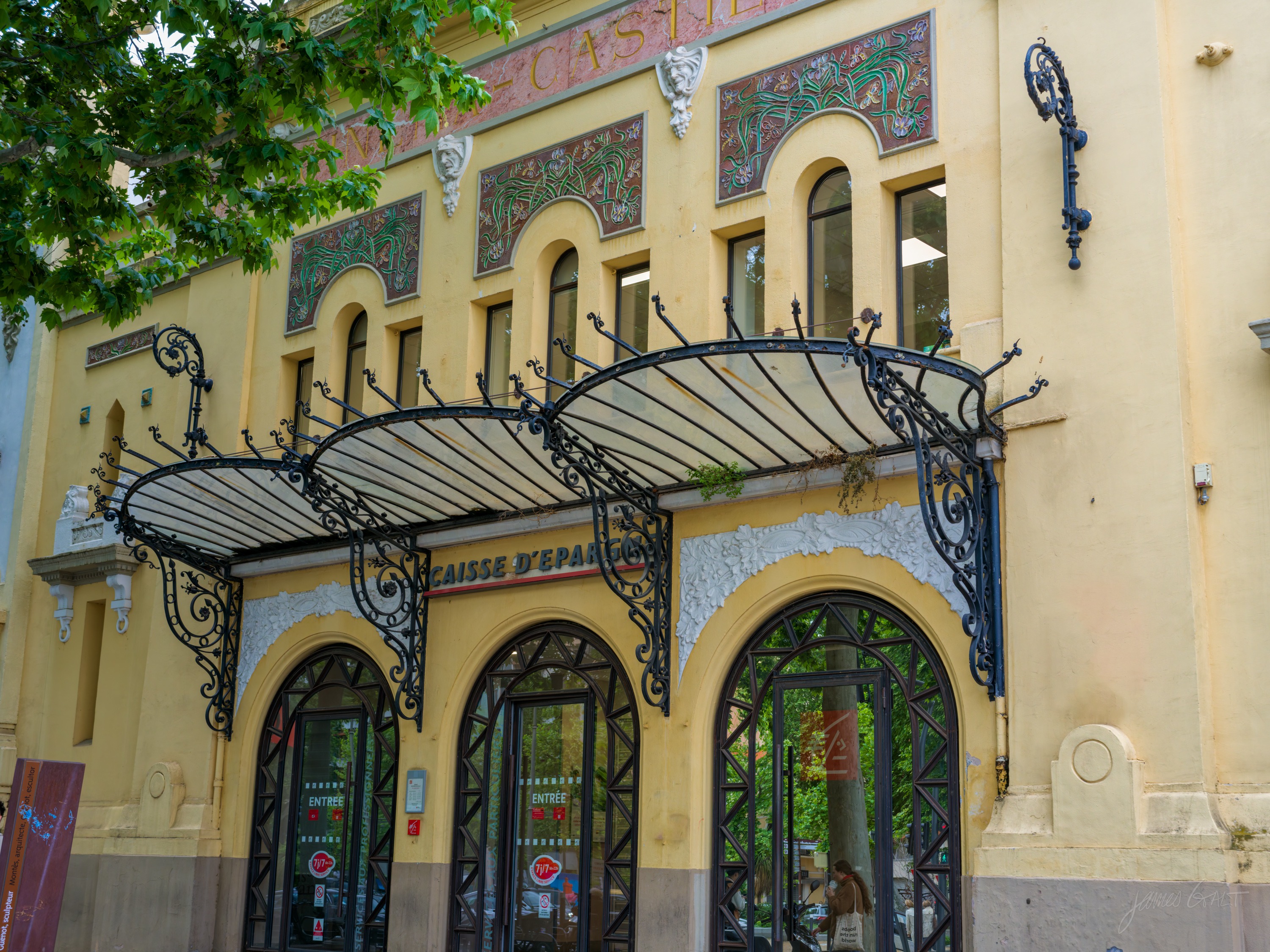 The facade of an old savings bank, three tall arched entrances shaded by curved wrought-iron and glass canopies, a painted frieze along the top.
