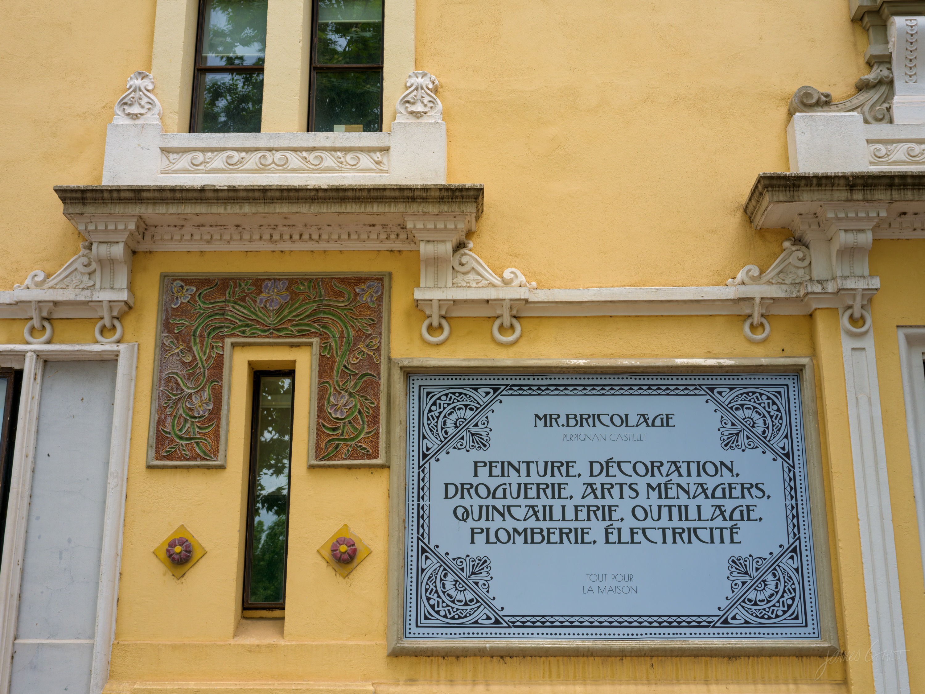 A yellow facade with a ceramic-tiled panel of lettering and floral borders set into the wall beside tall windows, corbels above.