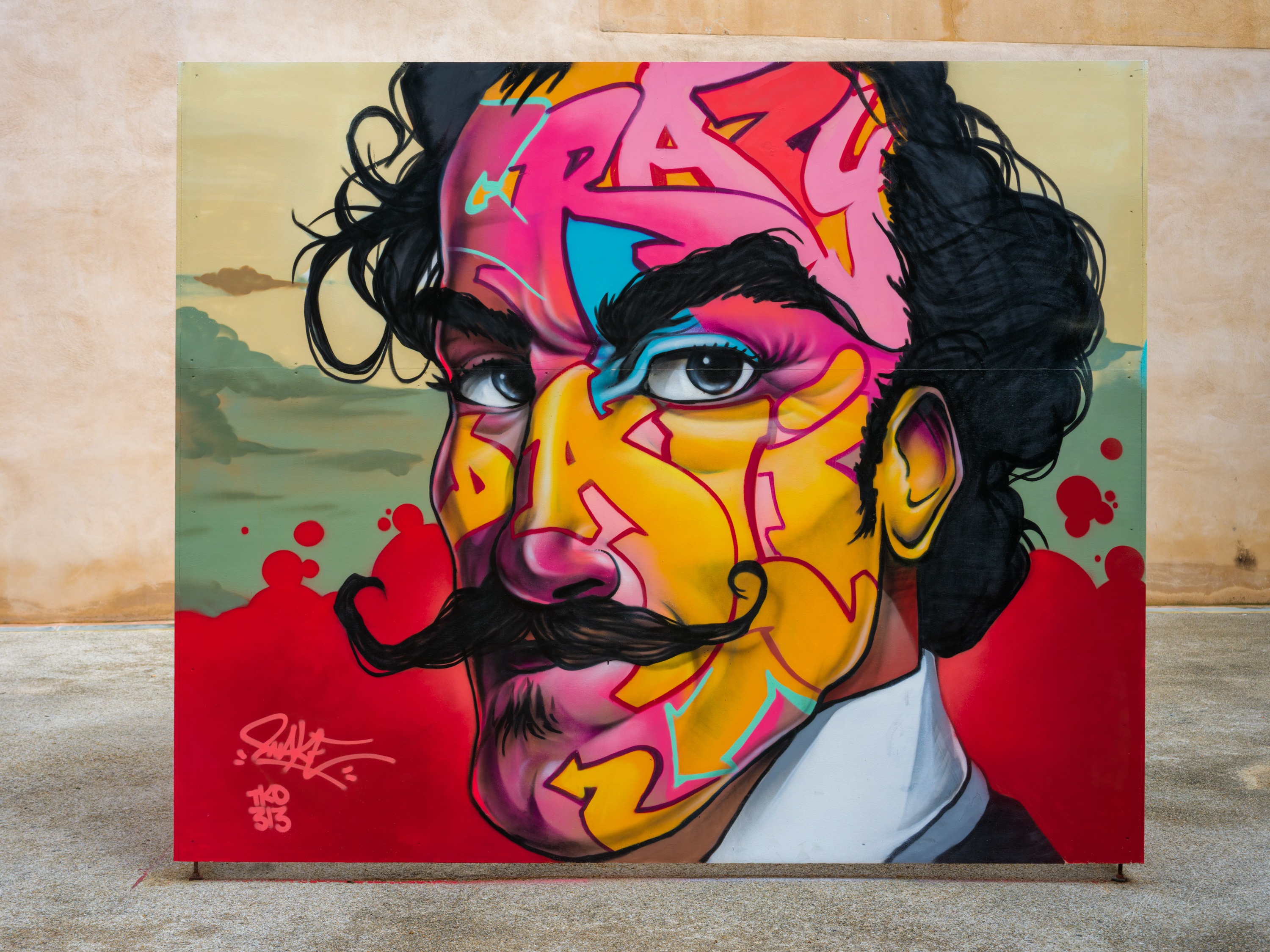 A large painted canvas propped against a wall: a stylised portrait of a moustached man in bright pink, yellow and blue graffiti style with the word CRAZY.