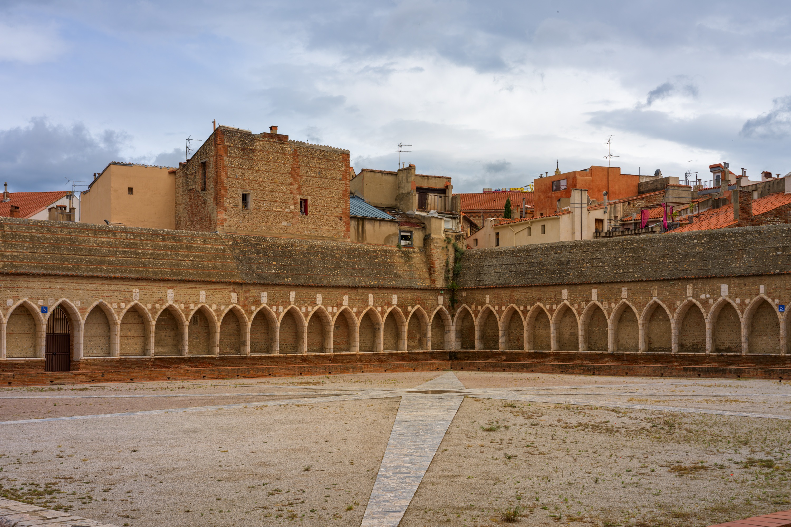 A vast open courtyard ringed by a continuous arcade of white marble Gothic arches, city rooftops rising behind one wall, bare gravel below.