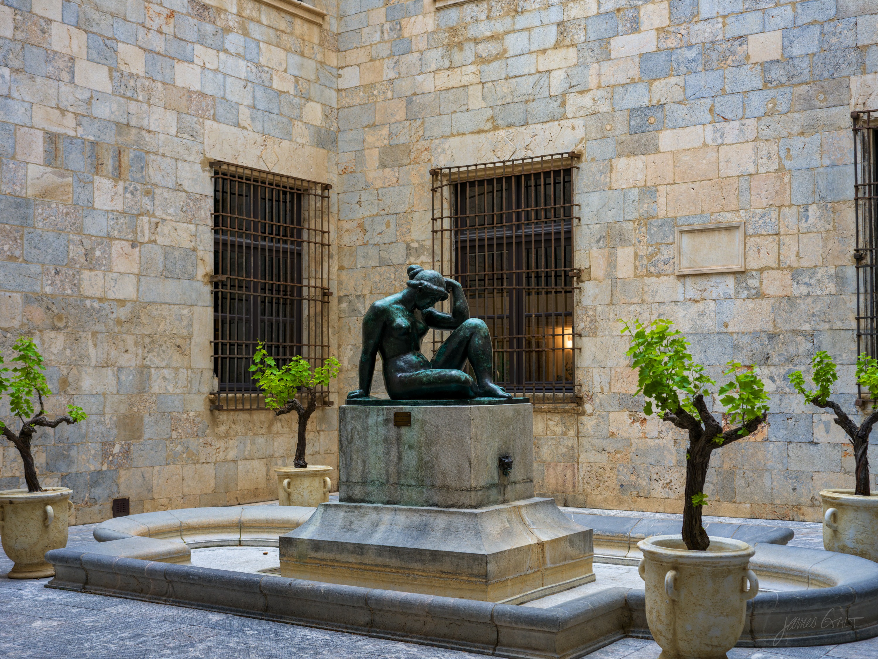 A green bronze of a seated nude woman, head resting on one raised arm, on a stone plinth in a fountain, potted orange trees against a barred stone wall behind.