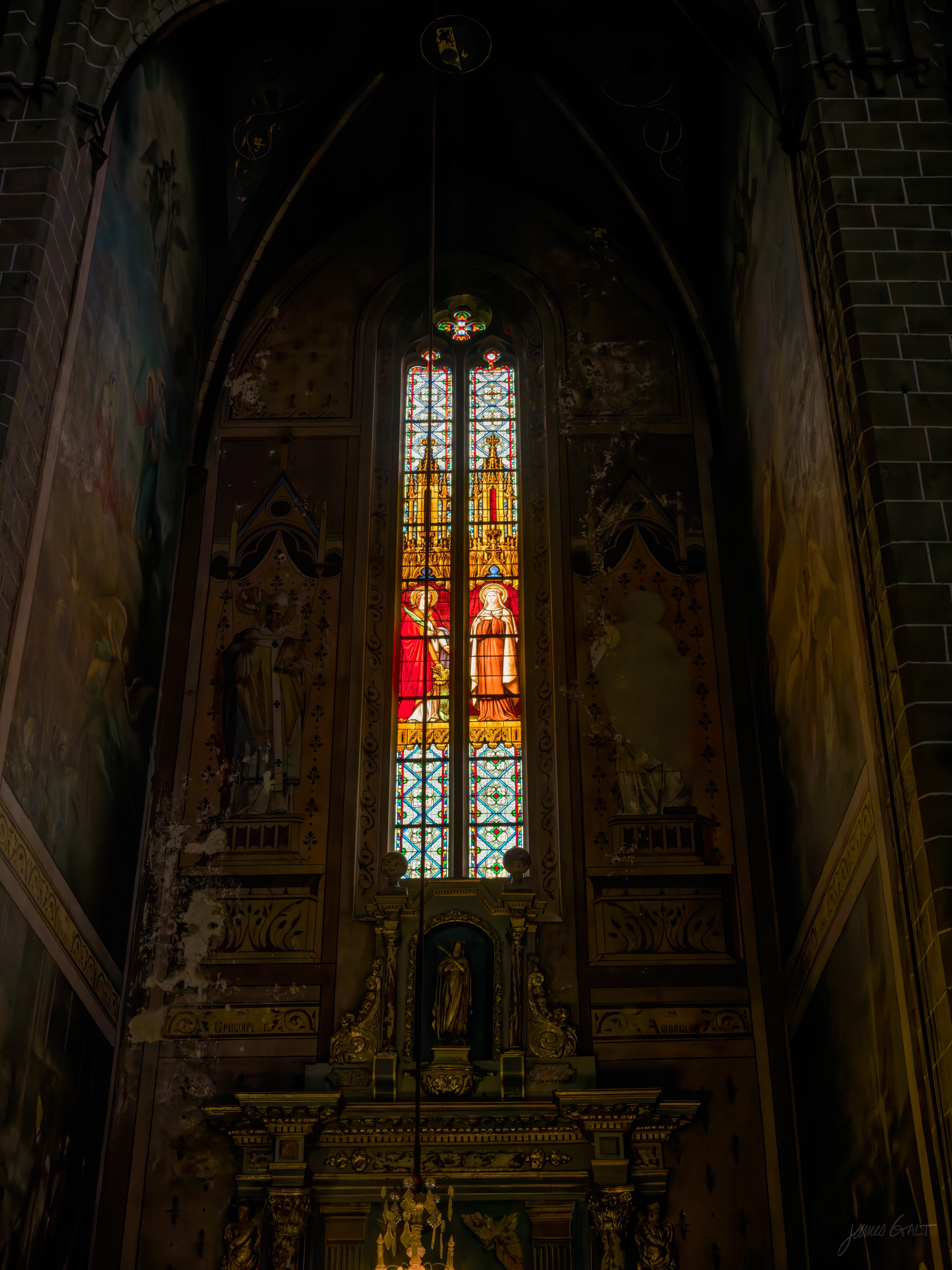 A tall stained-glass window glowing red and gold high in a dark cathedral, an ornate gilded altarpiece and painted walls below it, most of the frame in shadow.