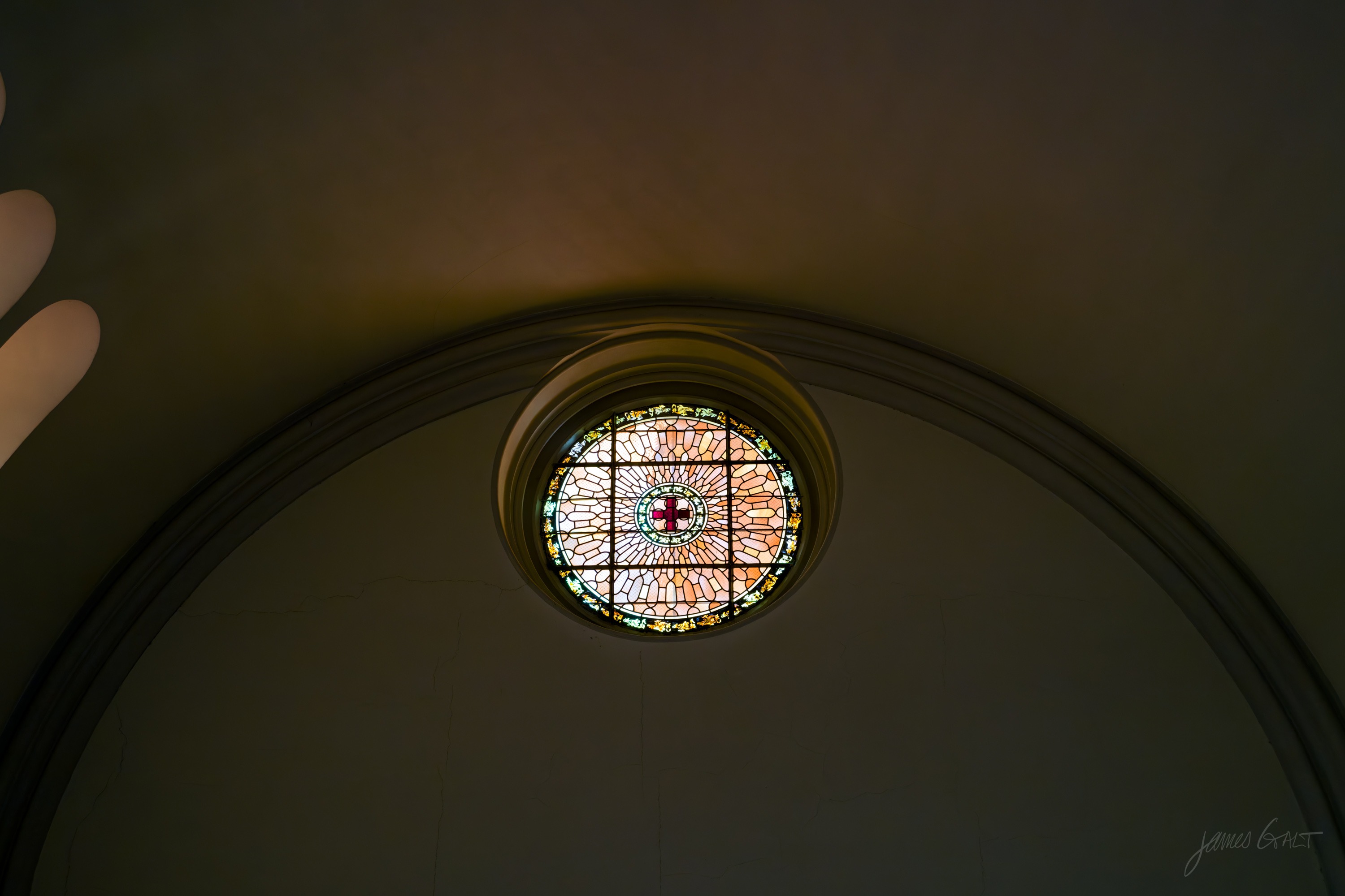 A small circular rose window of coloured glass set high in a dark plaster vault, the surrounding ceiling almost black, a hand just visible at the frame edge.