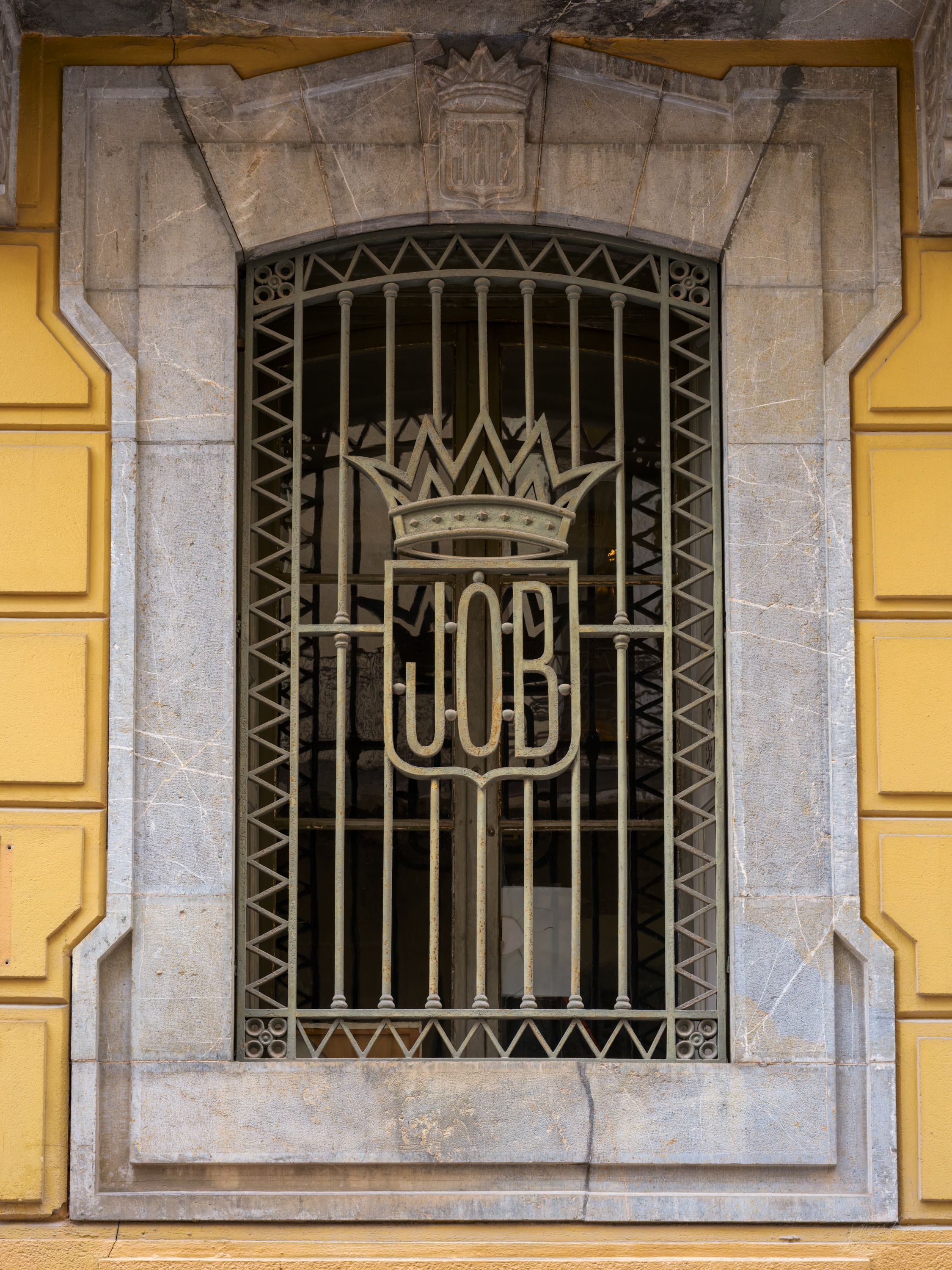 A tall arched window barred with an ornate iron grille worked into the monogram JOB beneath a crown, set in a stone surround on a yellow wall.