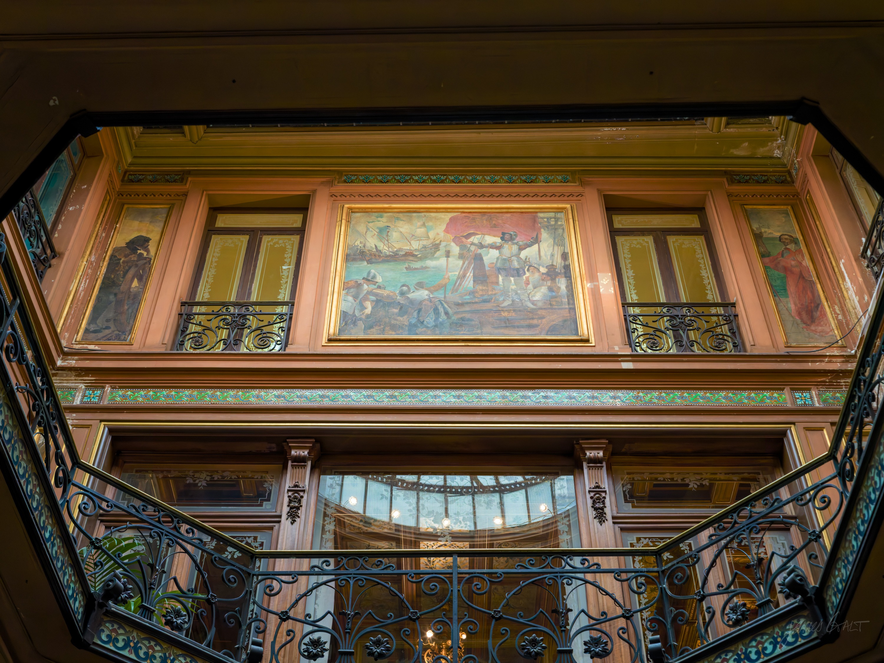 An ornate stairwell hall seen from below, a large painted mural above balconied landings with wrought-iron railings and gilded panels, warm light throughout.